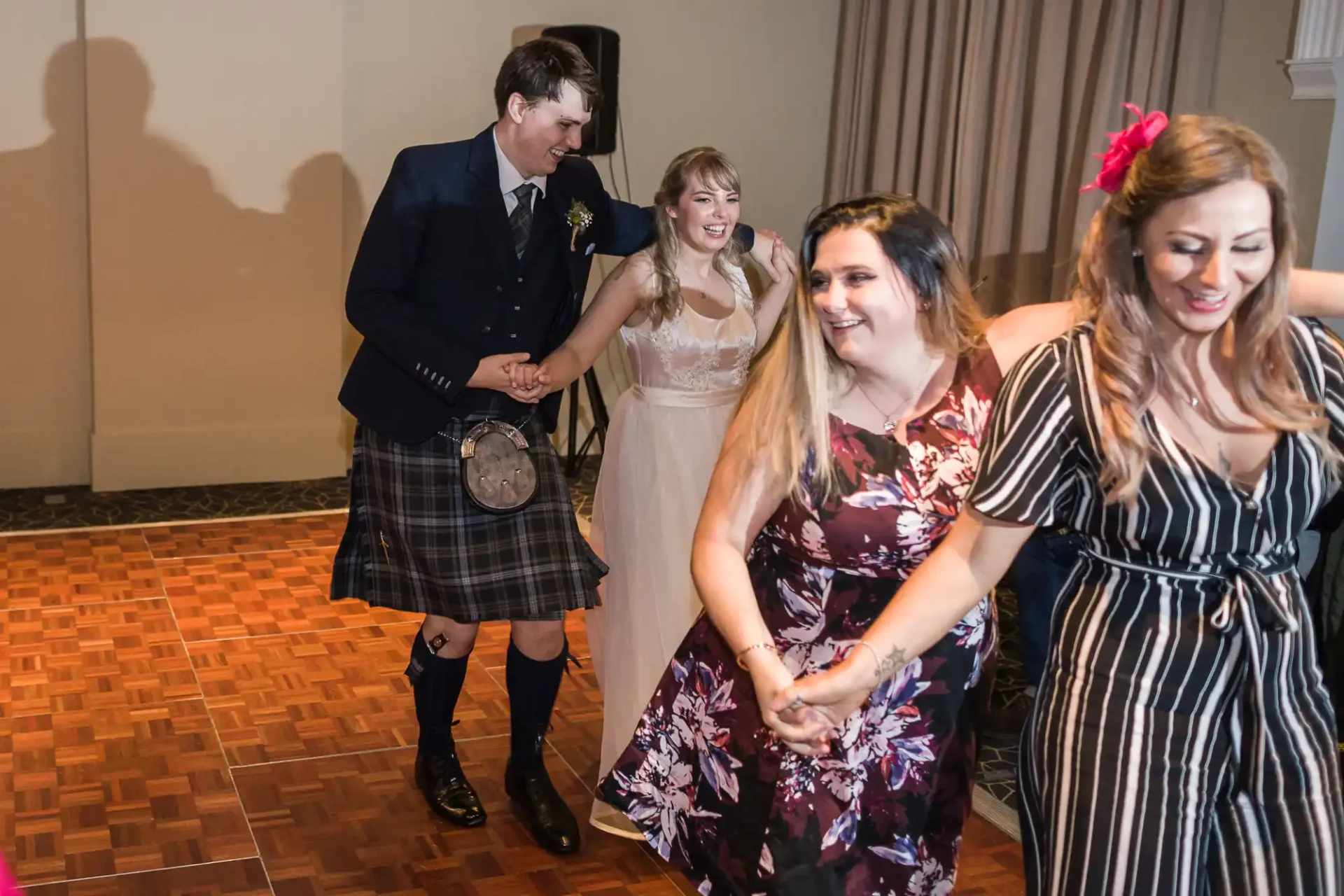 A group of people dancing joyfully at a wedding reception; one man wears a kilt and the women are dressed in floral and striped dresses.