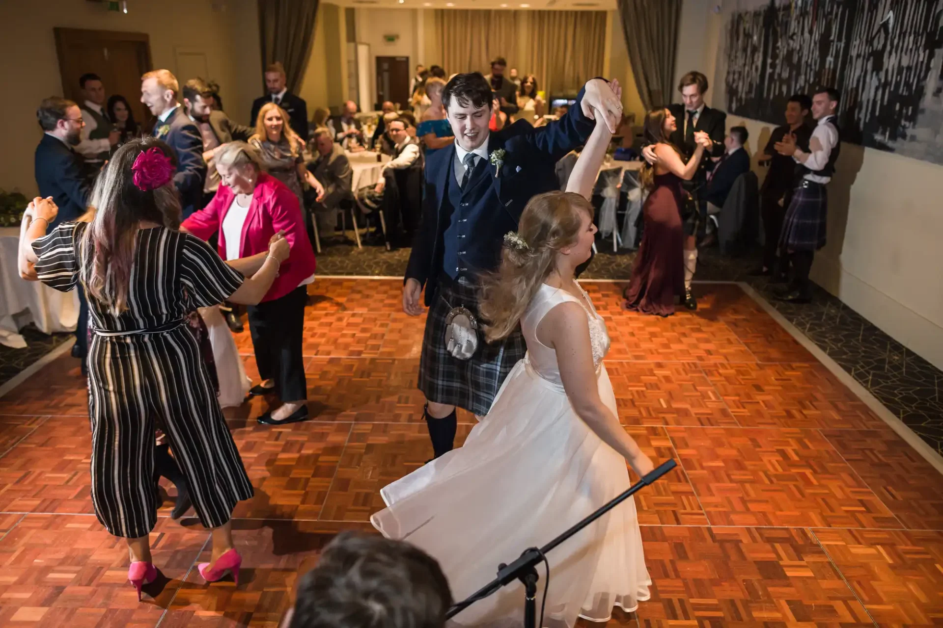 A bride and groom in traditional scottish attire dance joyfully with guests at their wedding reception.