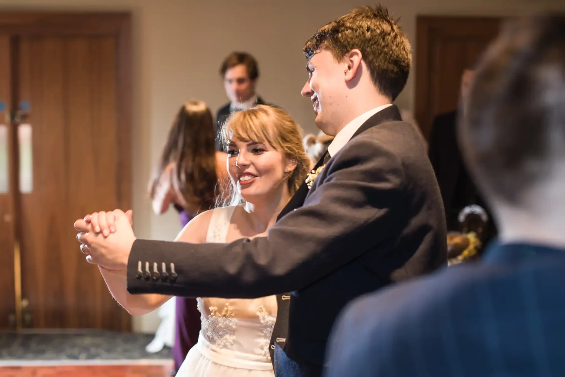A young couple dancing joyfully at a wedding reception, brightly smiling as they hold hands, surrounded by guests in soft focus.