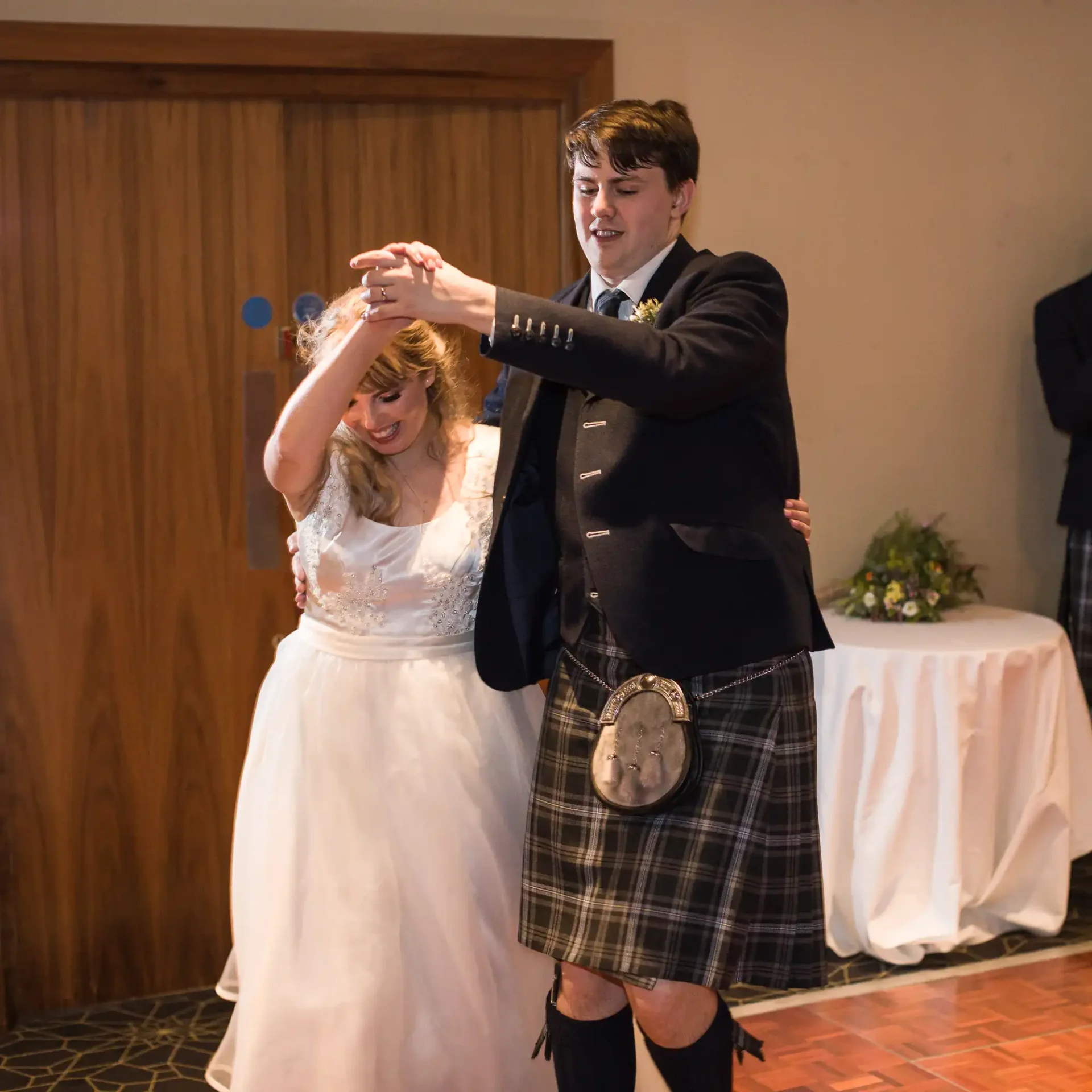 A bride and groom in traditional scottish attire dancing joyfully at their wedding reception.