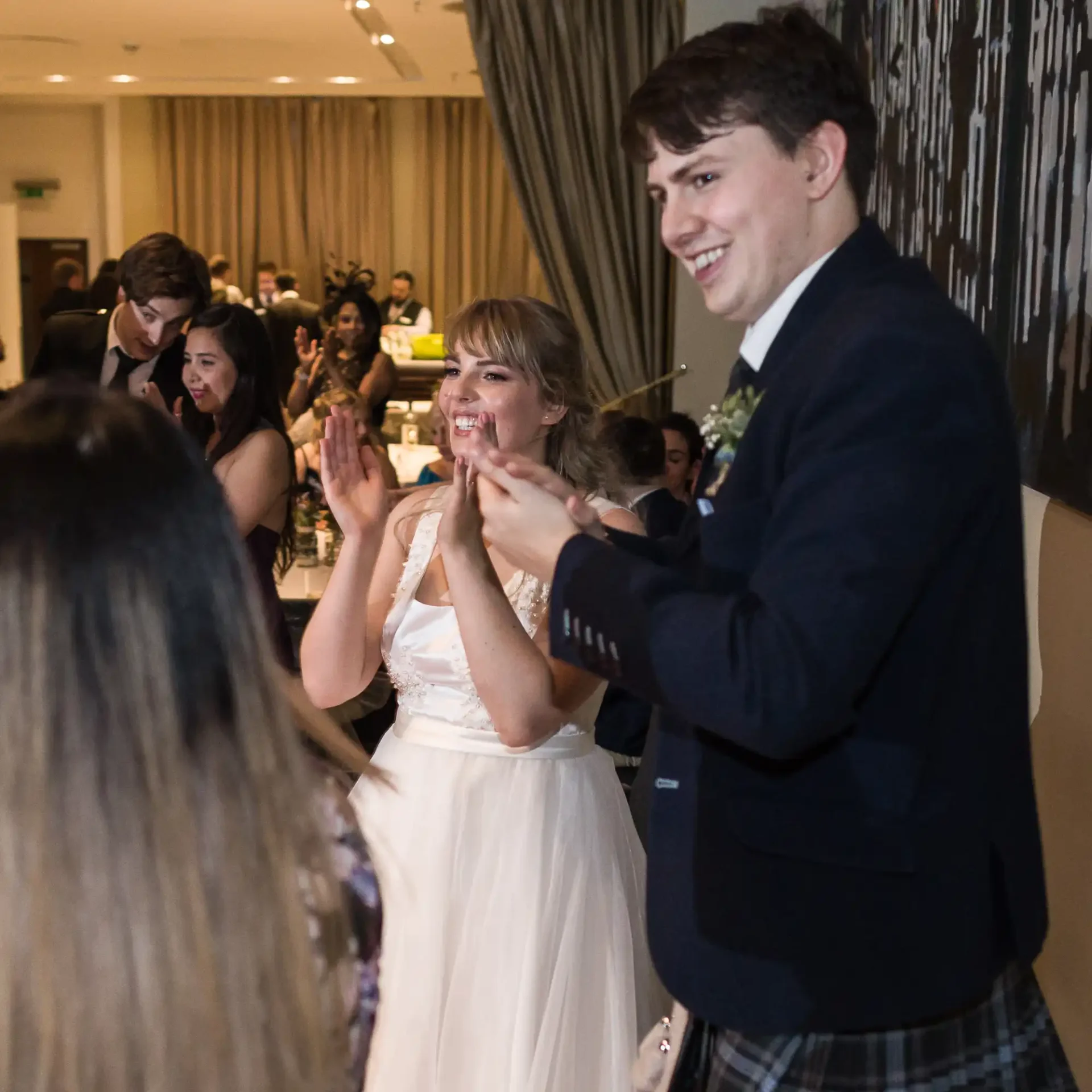 Bride in a white dress and groom in a kilt clap joyfully at a wedding reception surrounded by guests.