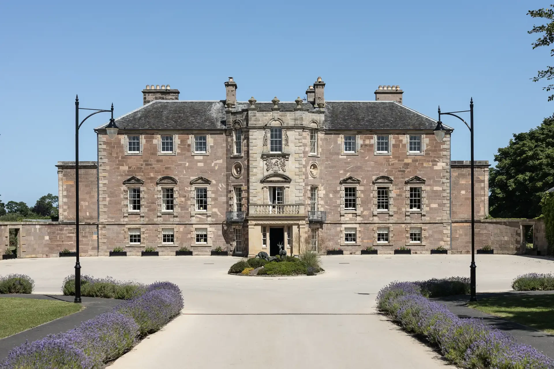 Archerfield House wedding photos: Front view of a grand, symmetrical stone mansion with a central entrance, flanked by lush lavender bushes and ornate lamp posts, under a clear blue sky.