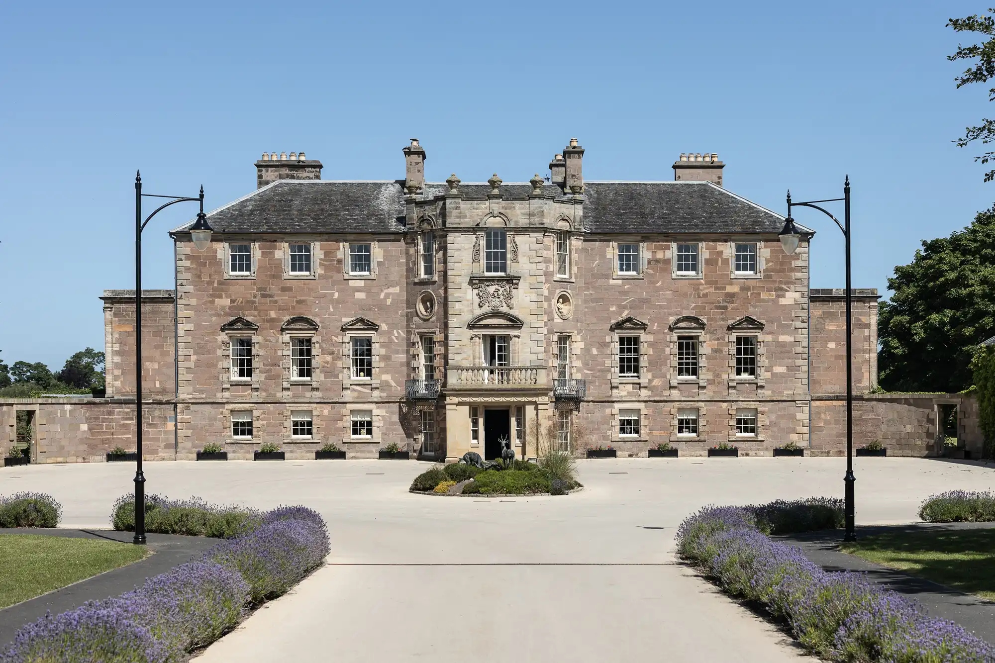 Front view of a grand, symmetrical stone mansion with a central entrance, flanked by lush lavender bushes and ornate lamp posts, under a clear blue sky.