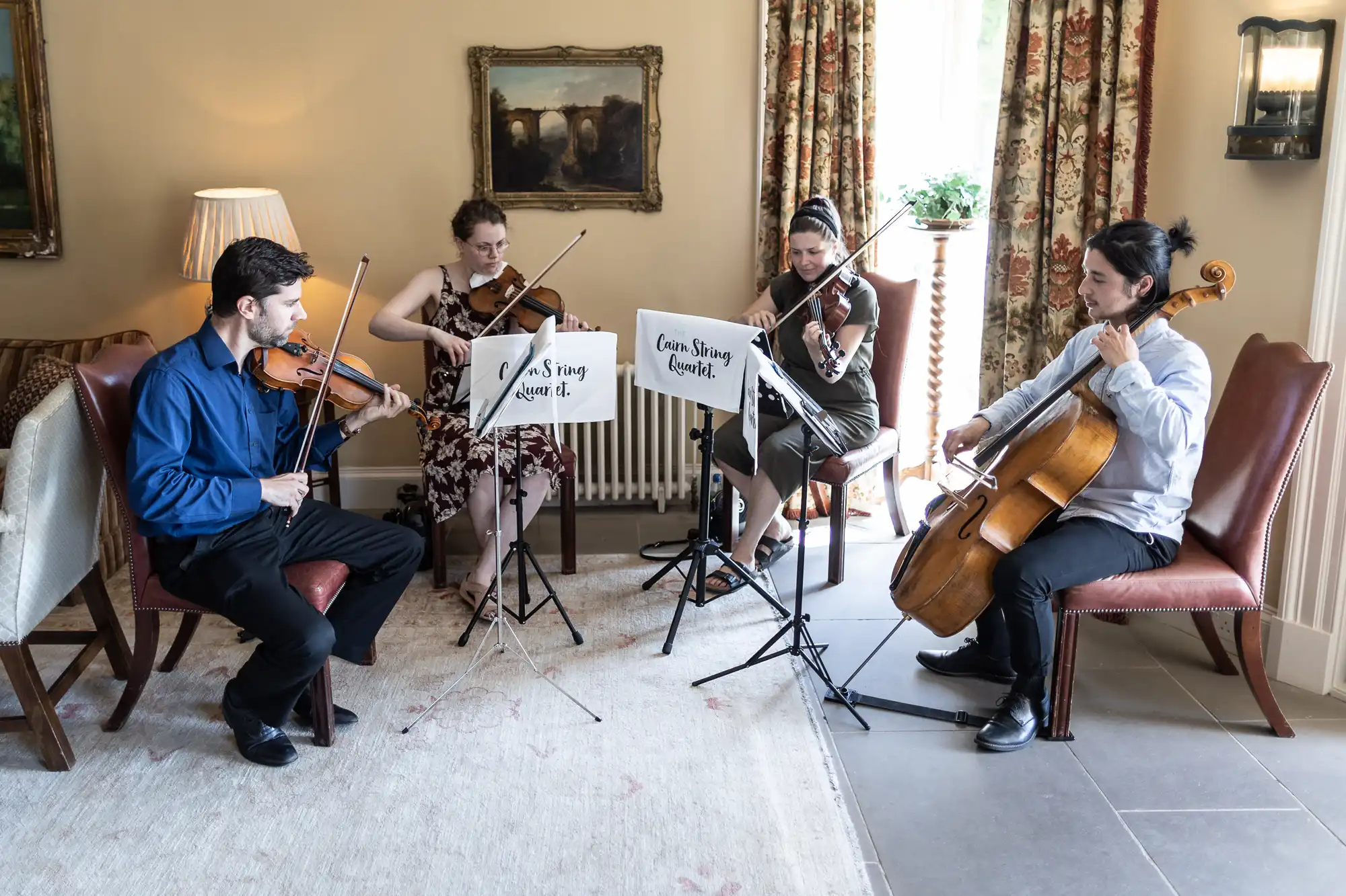 Four musicians, comprising two violinists, a violist, and a cellist, perform indoors, seated near a window, with a music stand labeled "cam. stg quartet.