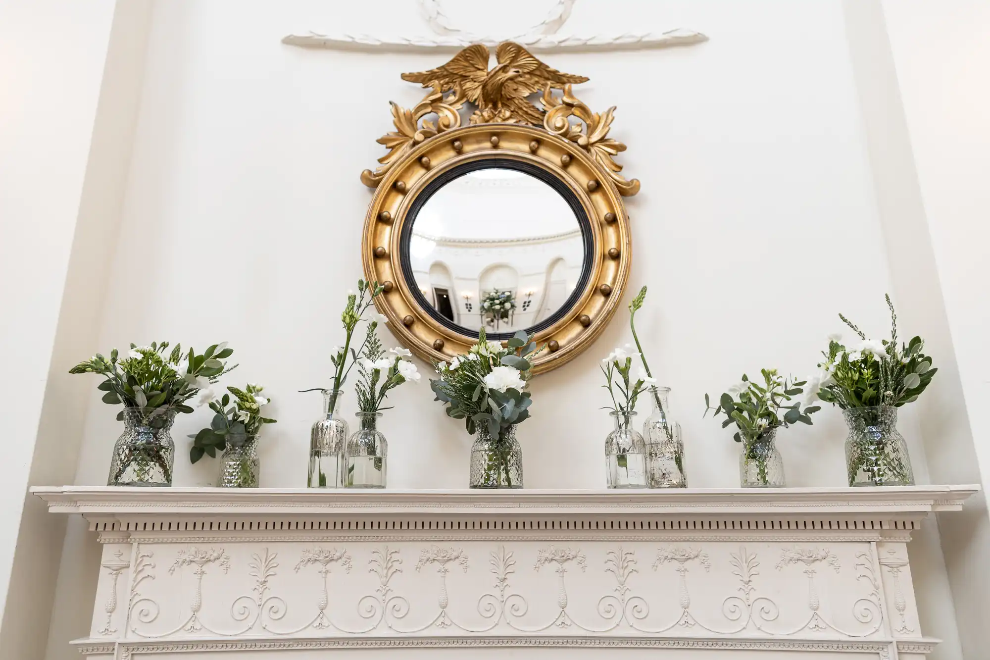 Decorative round gold mirror with an eagle motif, mounted on a white ornate mantelpiece flanked by green plants in glass vases.