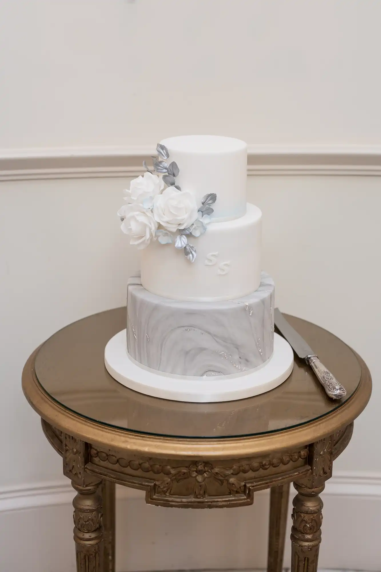 A three-tiered wedding cake with white and gray marbled design, decorated with white and silver flowers, displayed on a gold mirrored table with a cake knife.