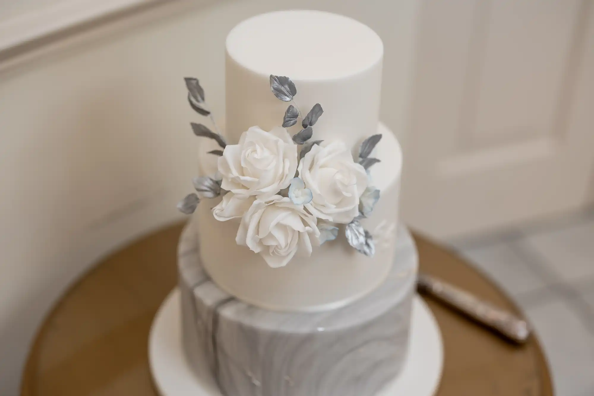 A two-tiered wedding cake with white and gray fondant, decorated with sugarcrafted white roses and gray leaves, displayed on a golden stand.