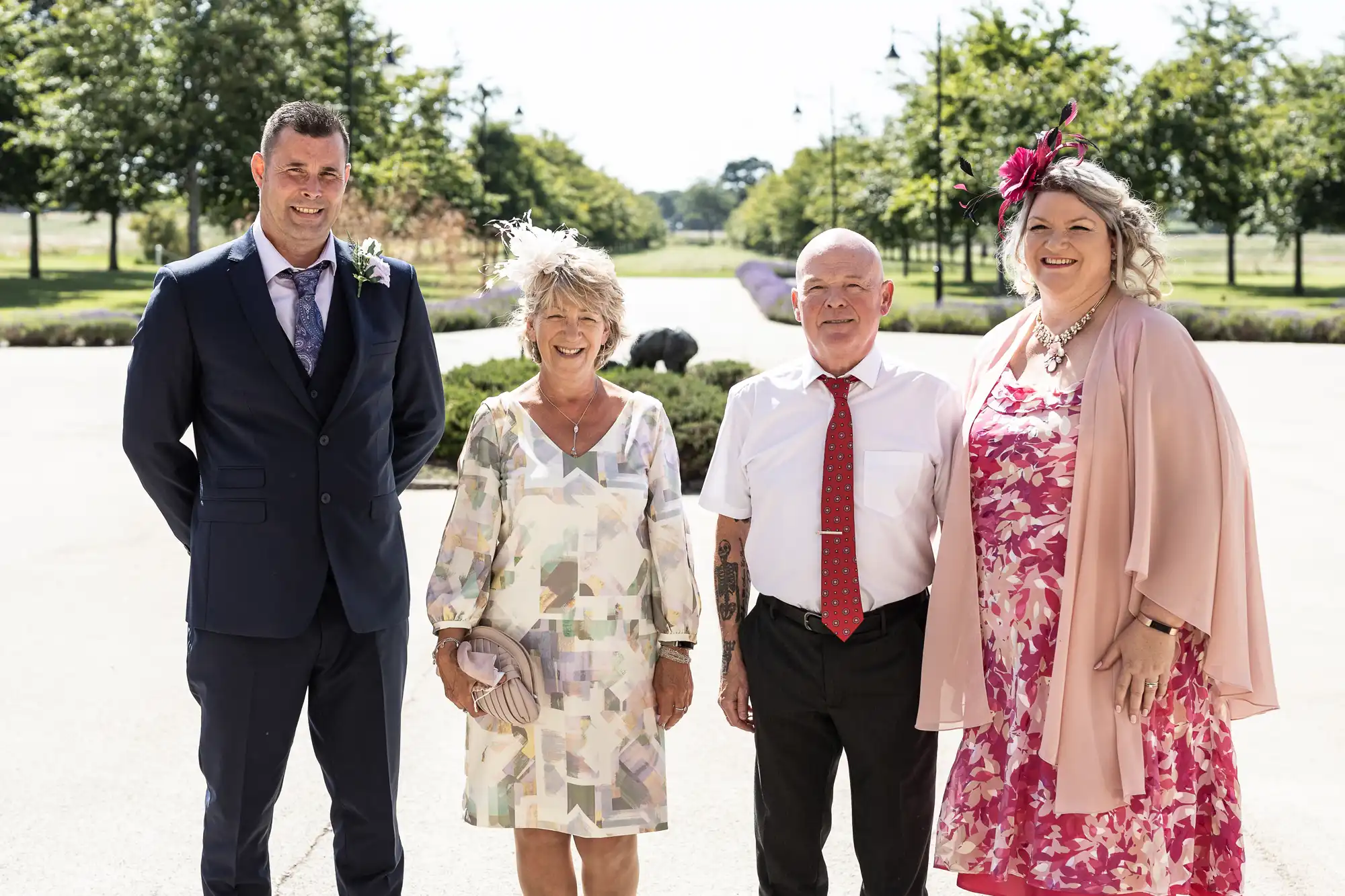 Four adults dressed in formal wear, smiling outside on a sunny day, with a park pathway and trees in the background.