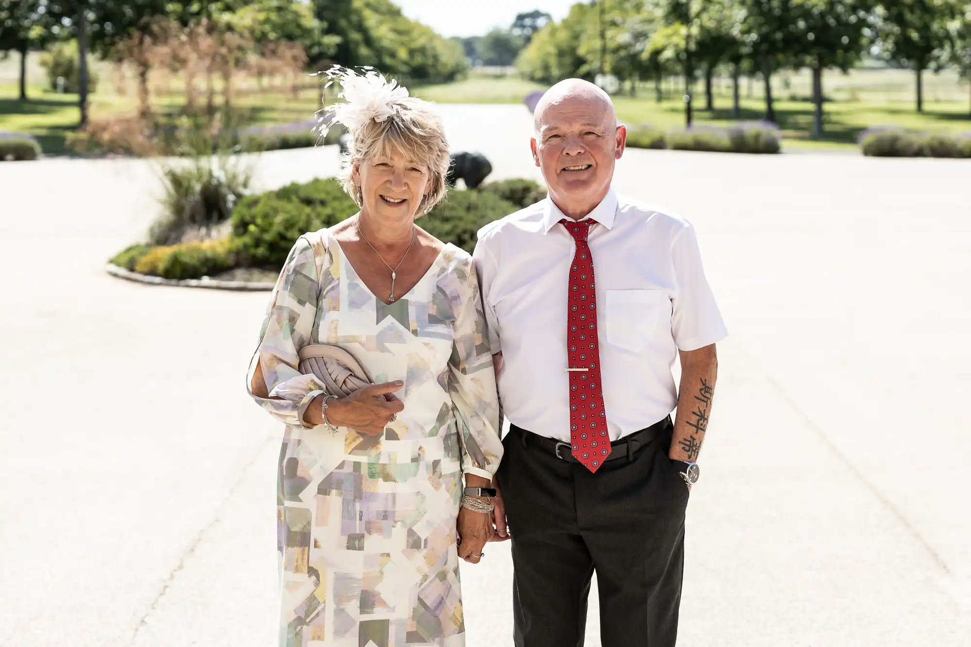 An elderly couple smiling and walking arm in arm outdoors, the woman in a floral dress and fascinator, the man in a white shirt and red tie.