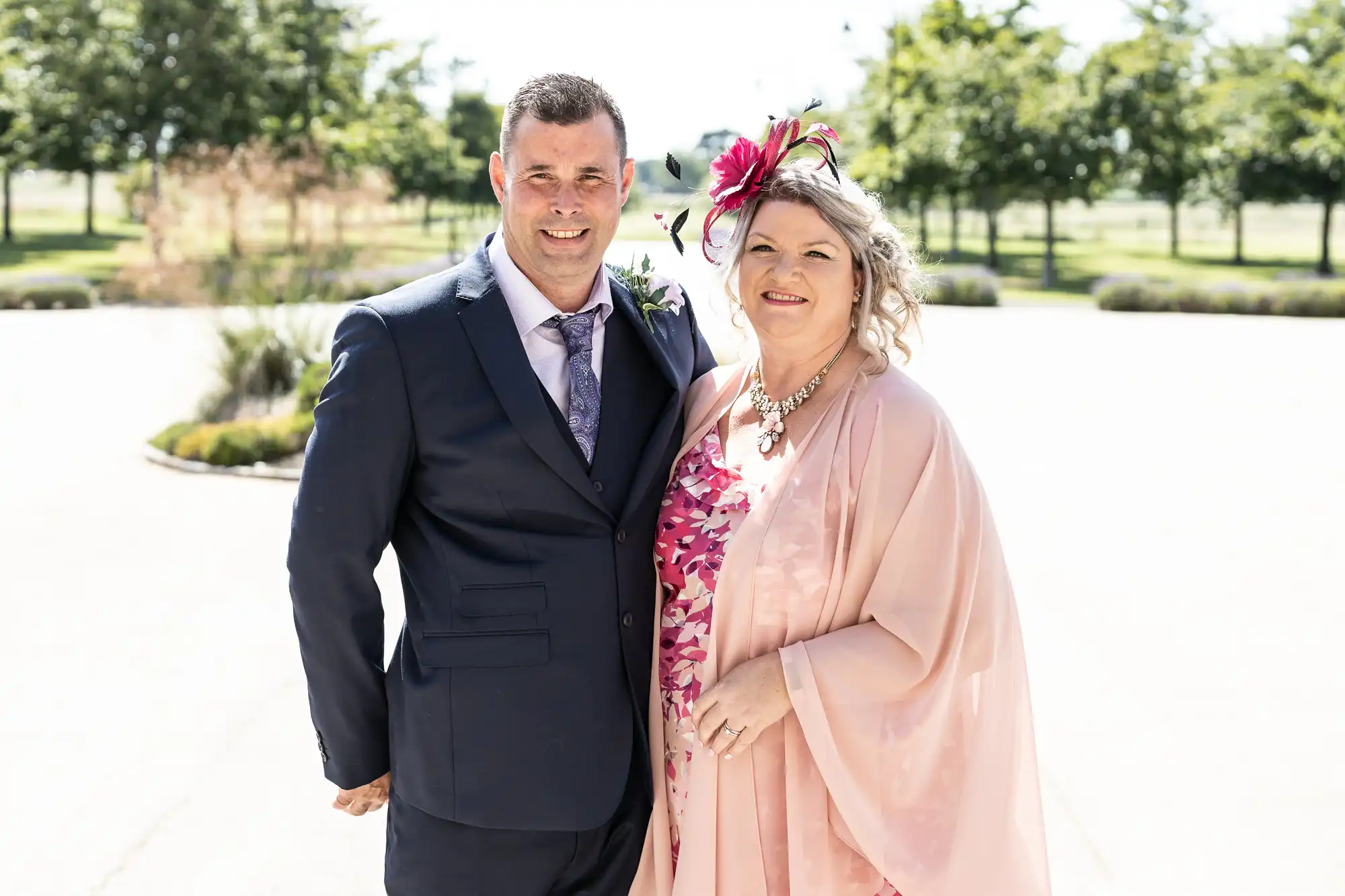 A man in a dark blue suit and a woman in a pink floral dress with a pink hat and shawl, standing together smiling on a sunny day with greenery behind them.