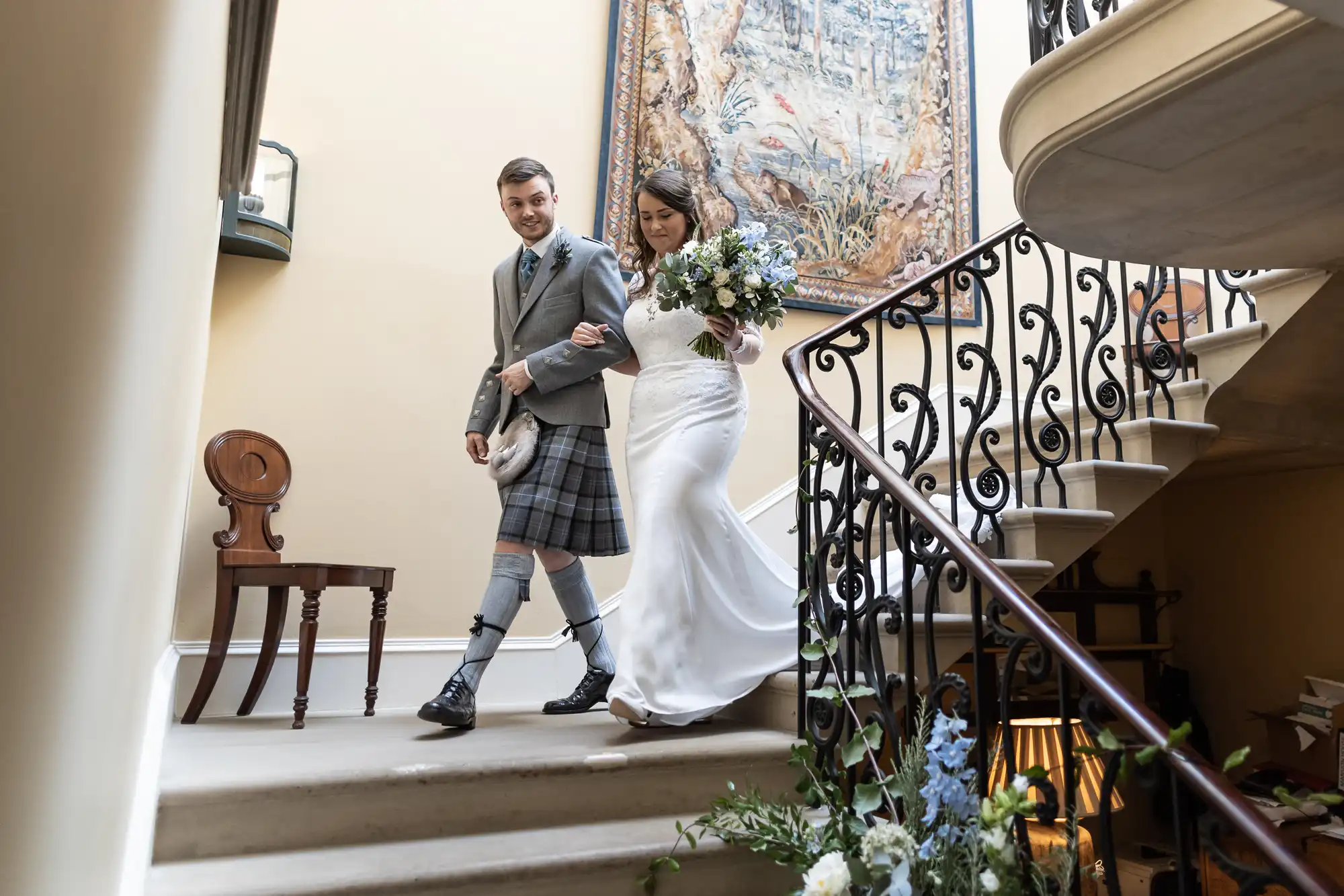 A bride and groom descending a staircase, the groom in a kilt and the bride in a long white dress, both smiling, with a floral bouquet in hand.