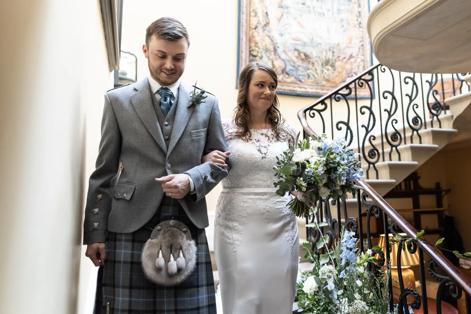 A bride and groom in wedding attire walking down a staircase, the groom in a kilt and the bride holding a bouquet.