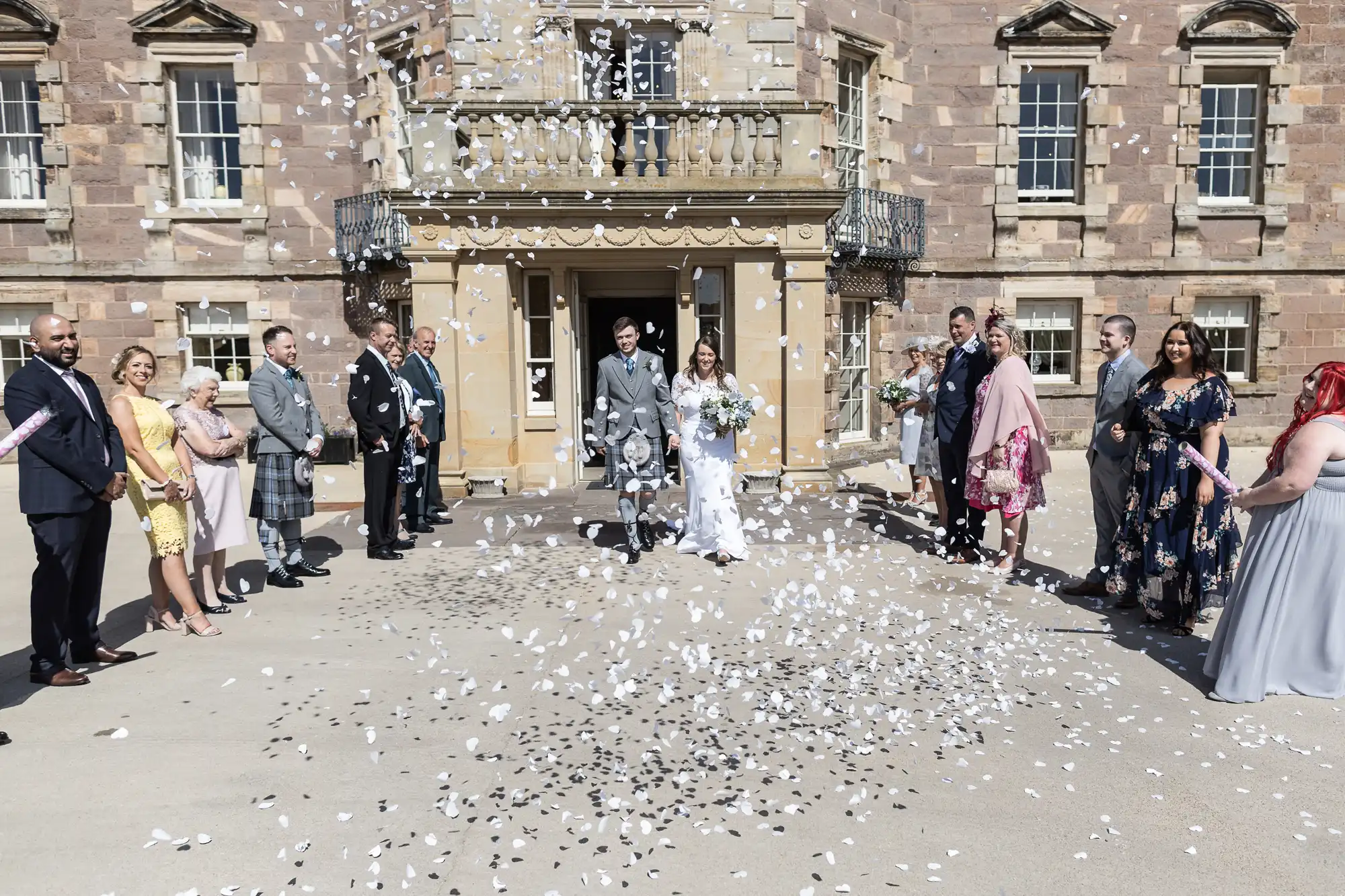 A newlywed couple walking out of a stone building, smiling as guests throw flower petals on a sunny day.