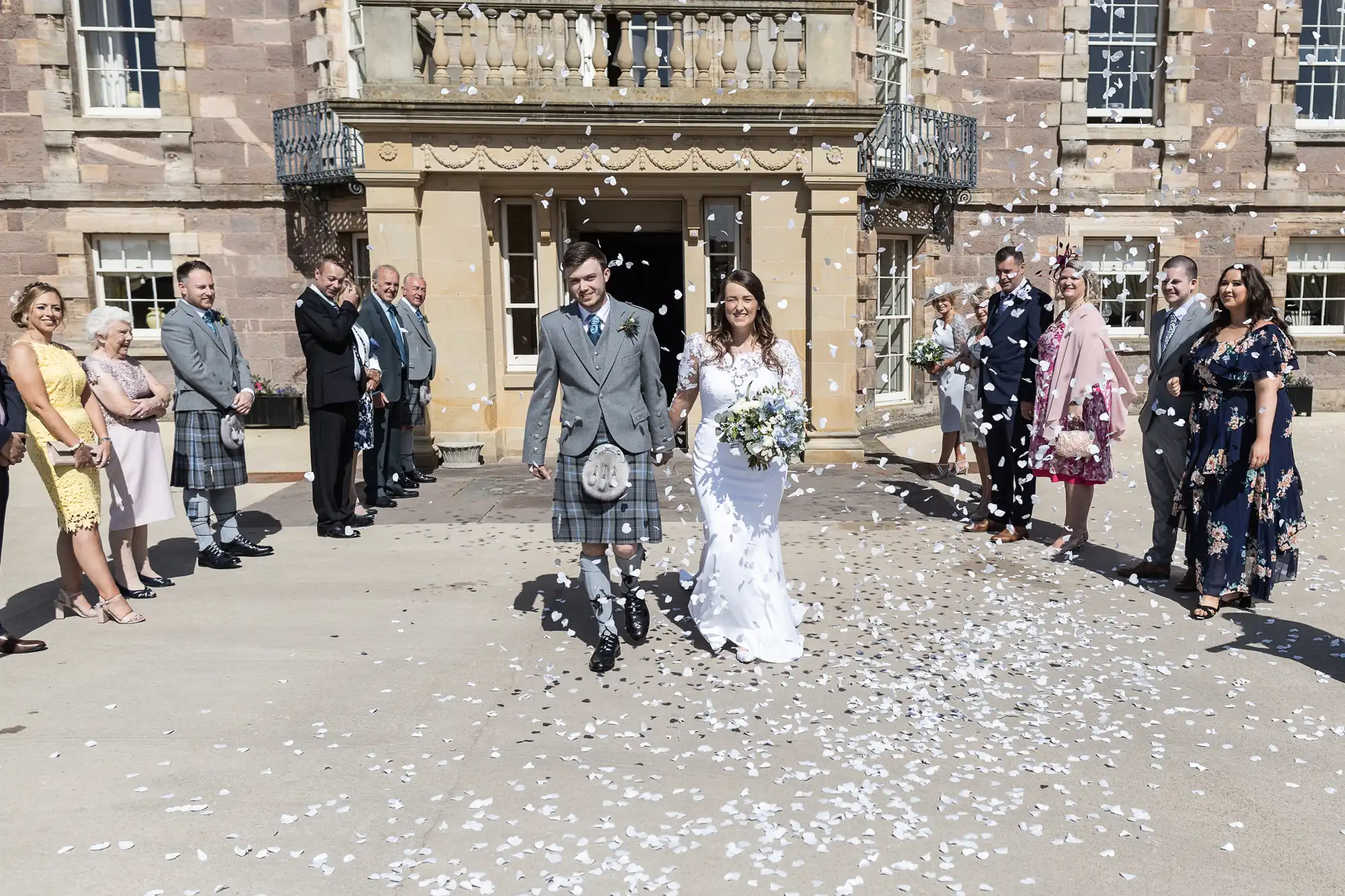 A bride and groom in traditional scottish attire exit a grand building, smiling as guests throw flower petals on a sunny day.