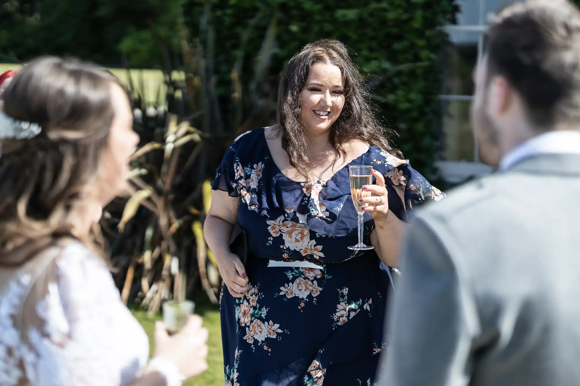 A woman in a floral dress holding a wine glass laughs with other guests at an outdoor gathering.