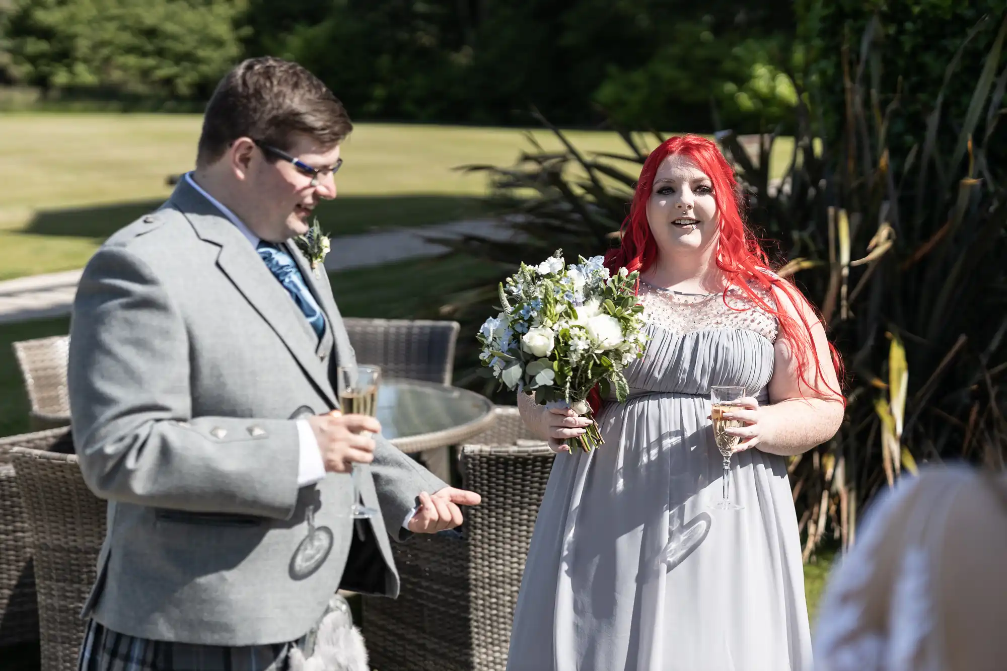 A bride with red hair in a white dress holding a bouquet converses with a man in a gray kilt and vest, both holding glasses, outdoors at a sunny wedding reception.