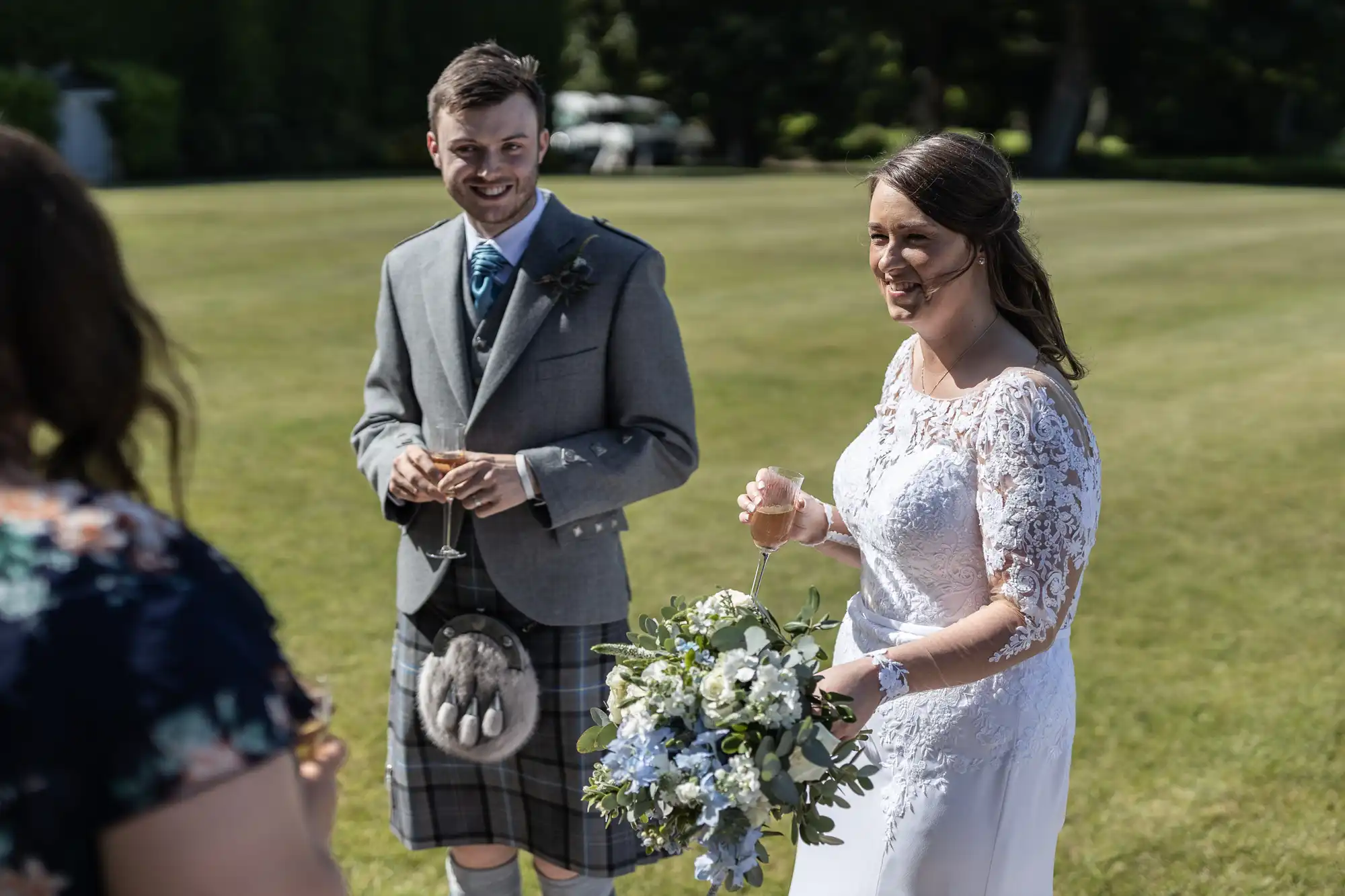 A bride and groom smiling outdoors at their wedding, with the groom in a kilt and the bride holding a bouquet.