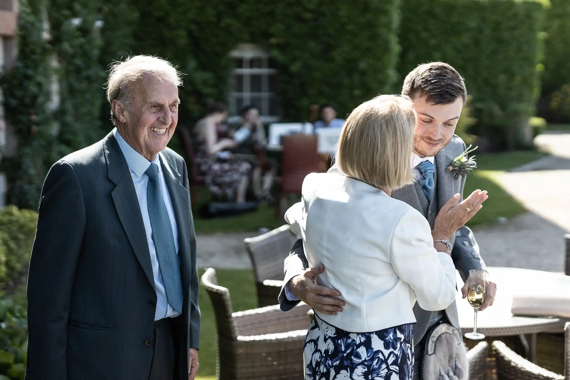 An elderly man smiles while looking on as a younger man in a suit hugs an older woman, all at an outdoor social gathering.