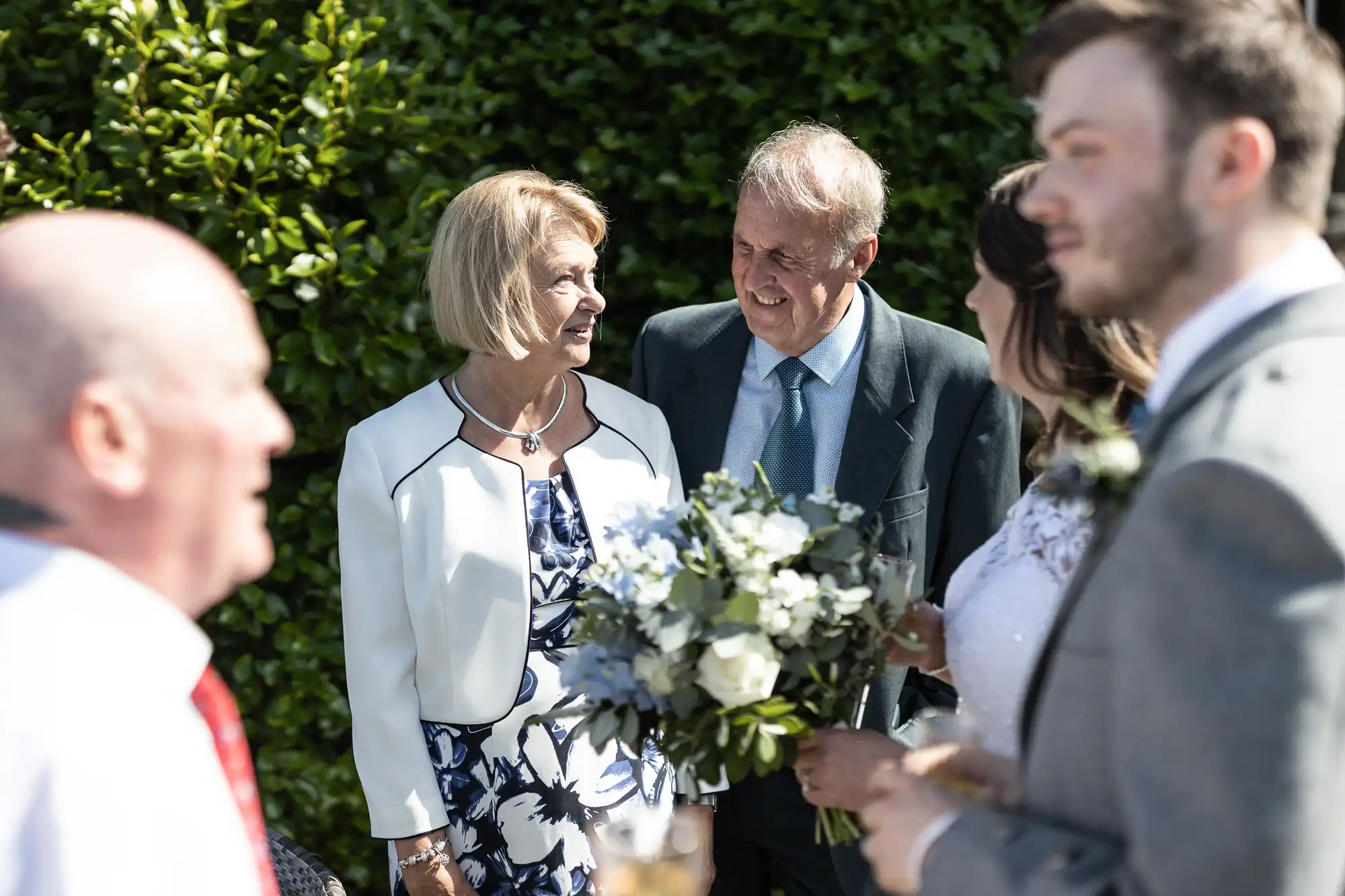 An elderly couple smiling and talking to guests at a wedding, surrounded by people holding flowers in a sunny garden setting.