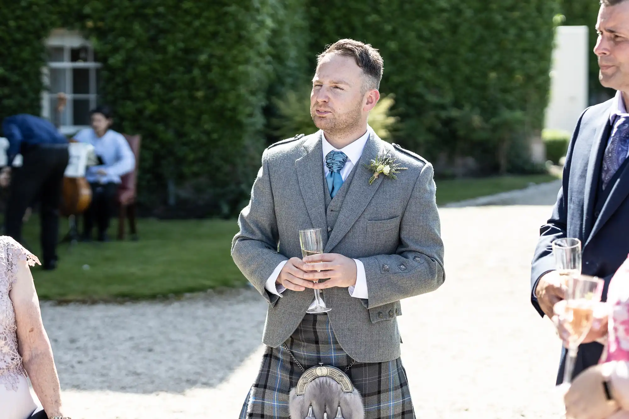 A man in a traditional scottish kilt and tweed jacket holds a champagne glass at a sunny outdoor wedding reception.