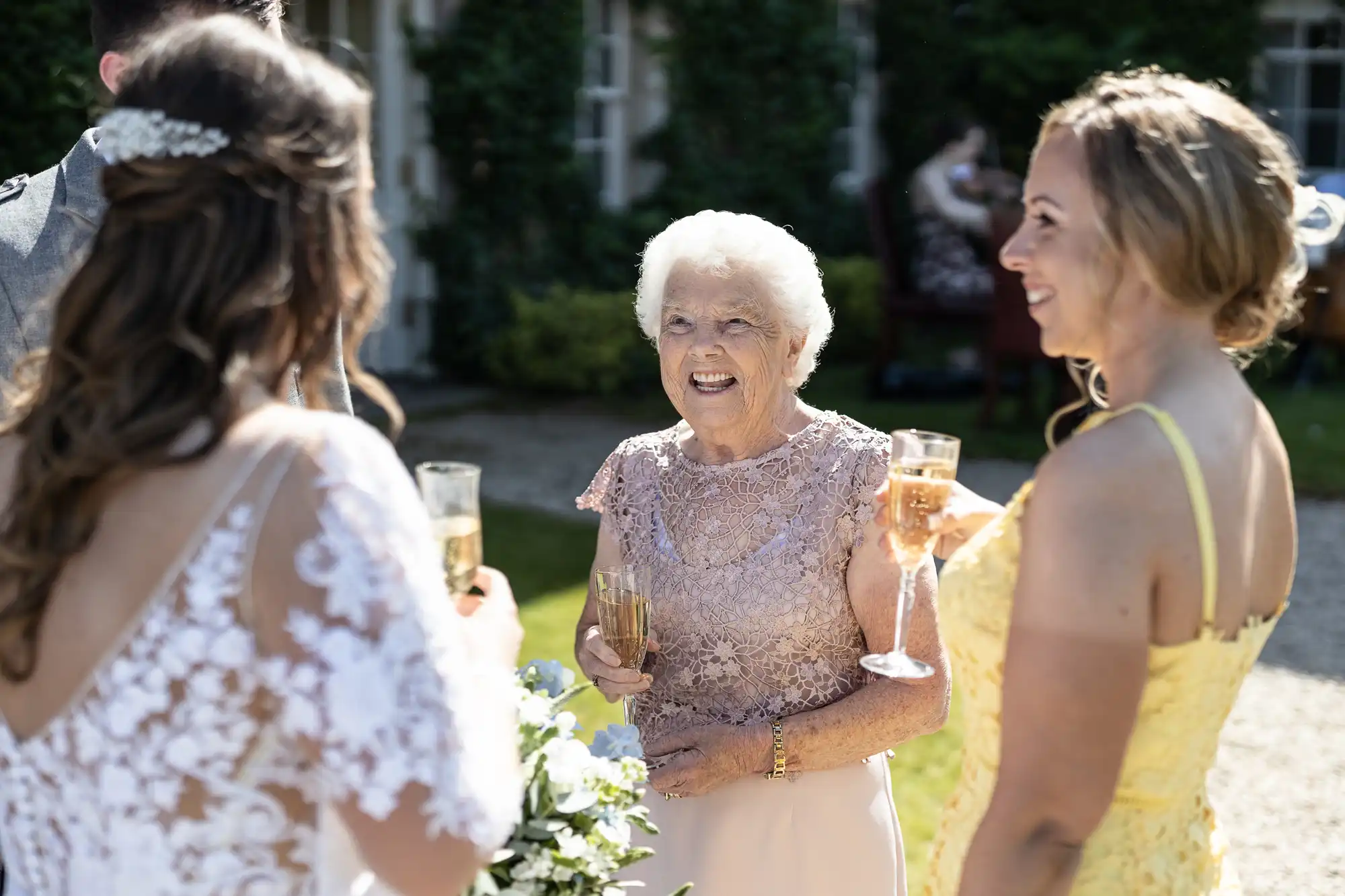 An elderly woman in a pink lace dress smiles joyfully at a bride and another guest, all holding champagne glasses at a sunny, outdoor wedding celebration.