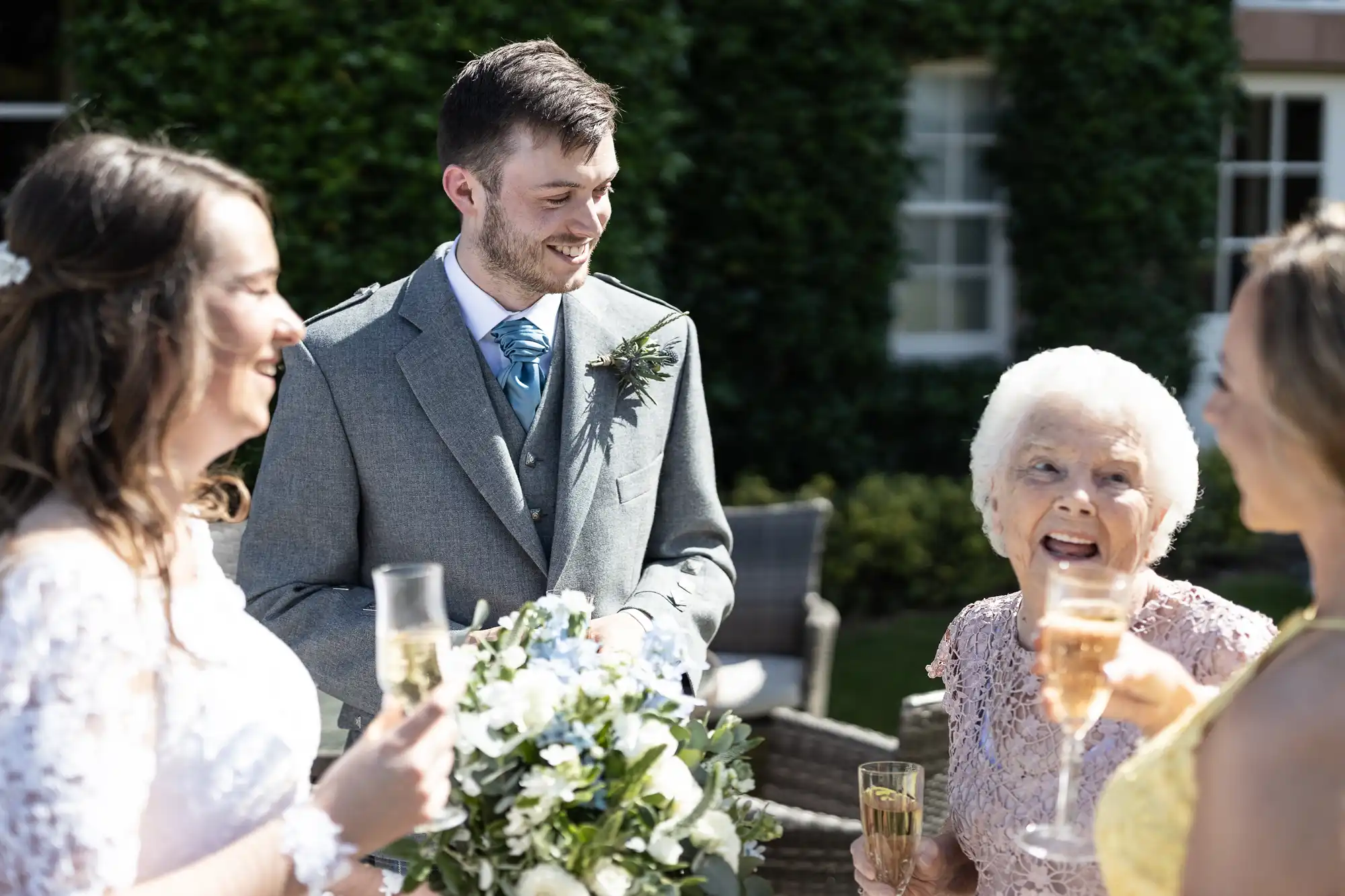 A groom smiling and chatting with three women during a sunny outdoor wedding reception, with champagne glasses in hand.