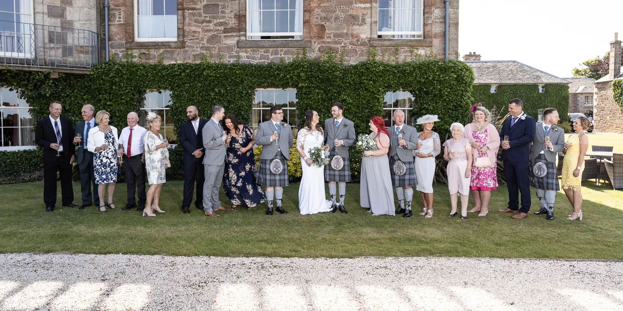 A group of wedding guests holding hats stands in front of a stone building on a sunny day, awaiting a formal event.