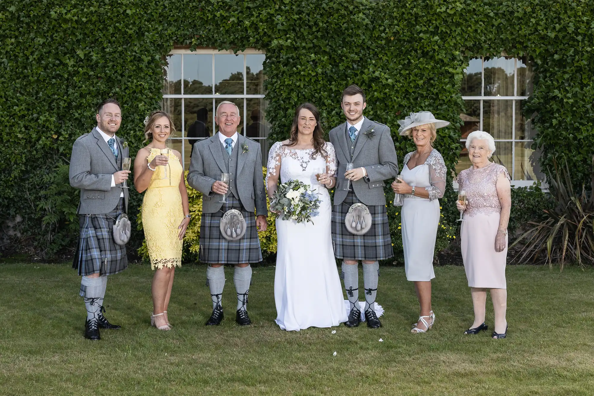 A family dressed in formal attire with kilts poses for a photo at a wedding, holding champagne glasses, in front of a hedge.