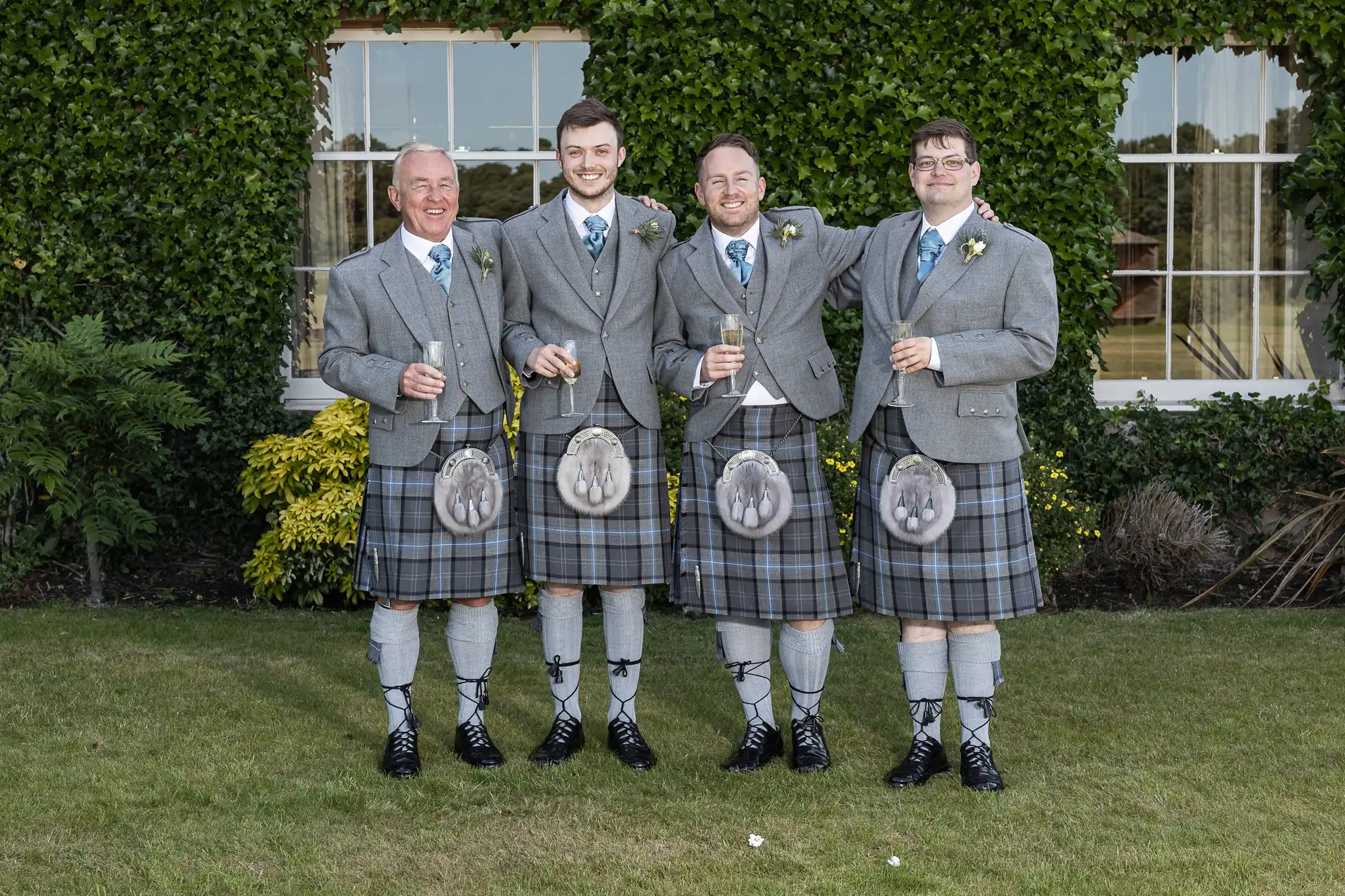 Four men in traditional tartan kilts and blazers, smiling and holding whisky glasses, standing on a lawn with a hedge backdrop.