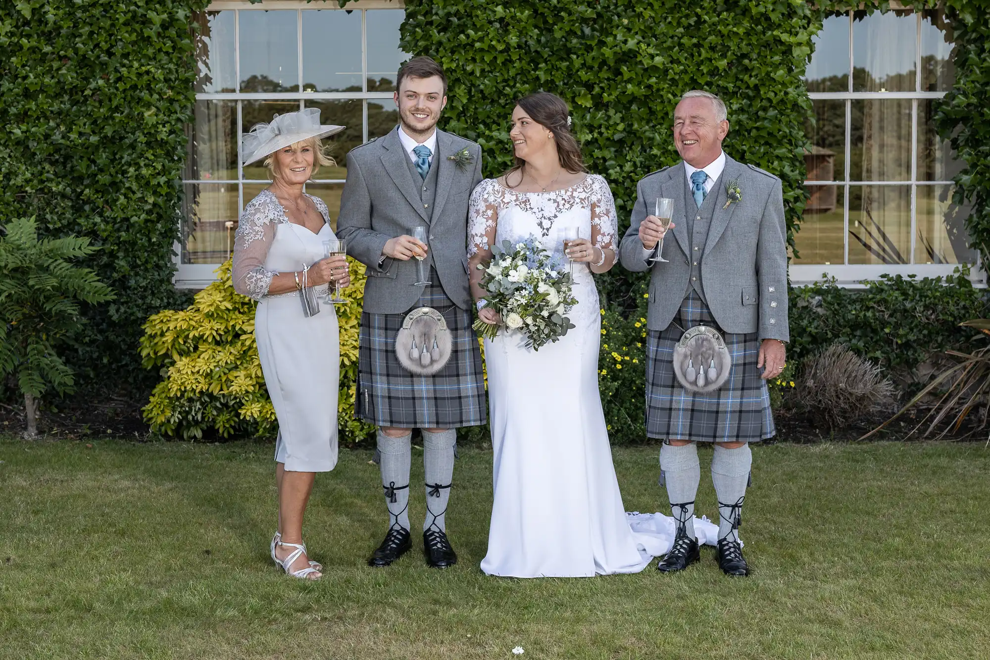 A wedding photo featuring a bride and groom with two older adults, all smiling; men in kilts and women in dresses, standing on a lawn before a building.