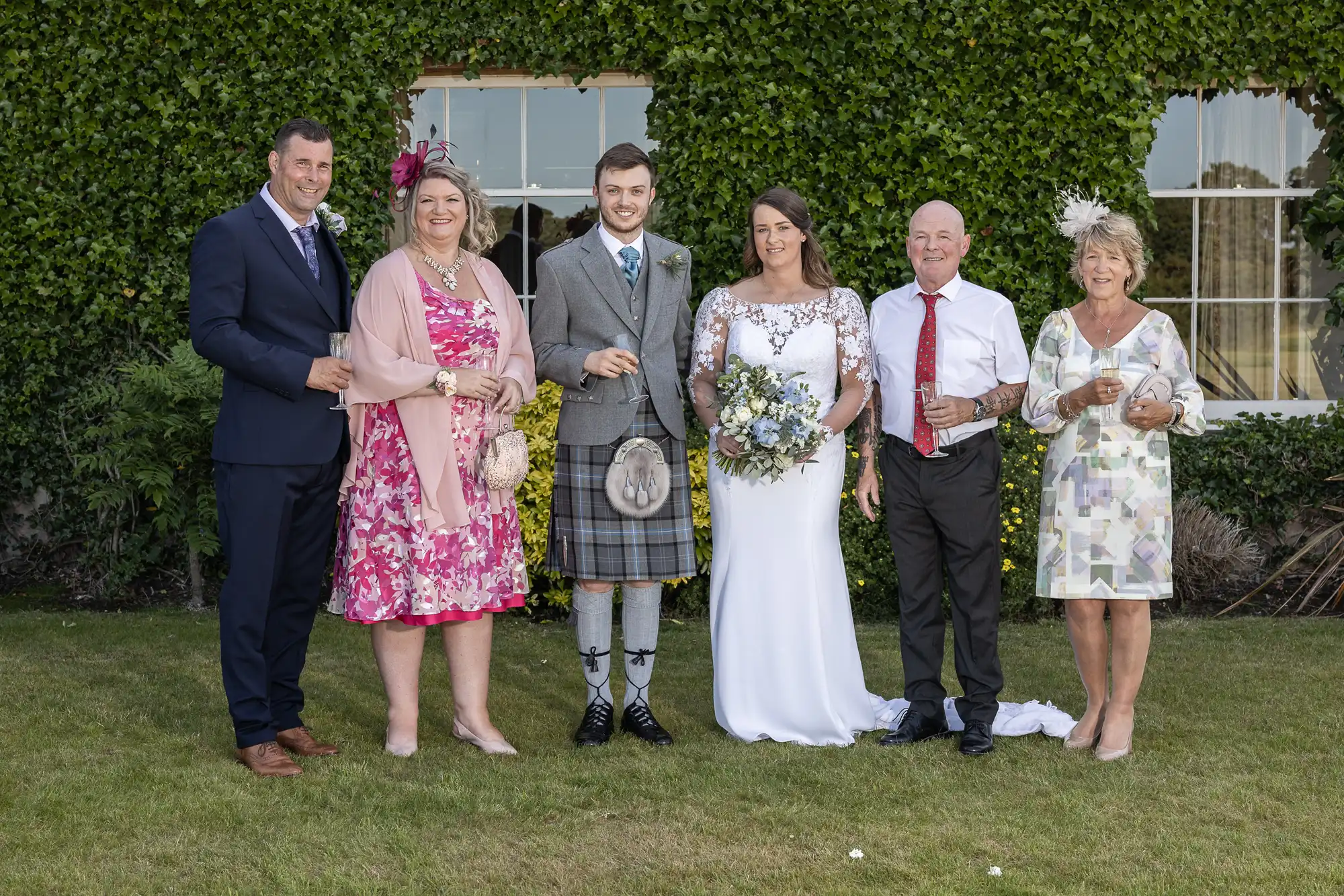 A wedding group with six people posing on a lawn; the groom is in a kilt, and the bride is in a white dress.
