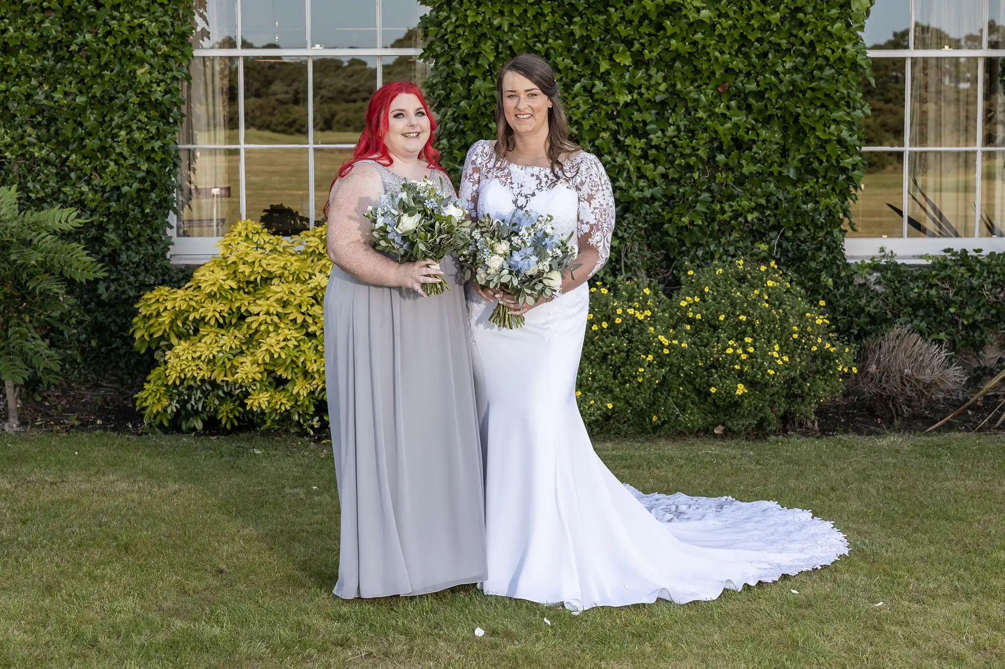 Two women, one in a white wedding dress and the other in a gray gown, holding bouquets and smiling in a garden with a building in the background.