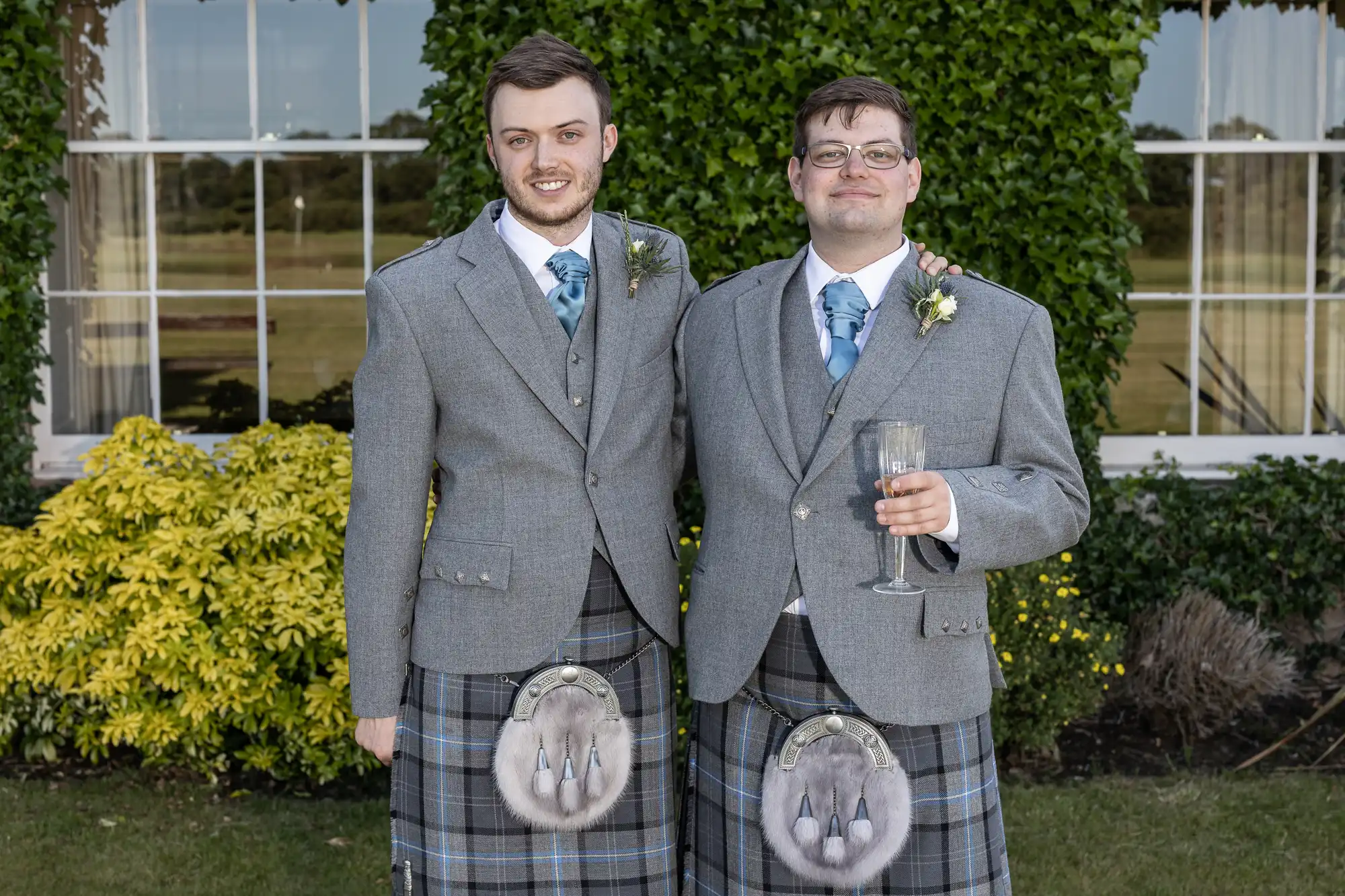 Two men in traditional scottish attire, including kilts and tweed jackets, smiling and holding champagne glasses, standing outdoors at a formal event.