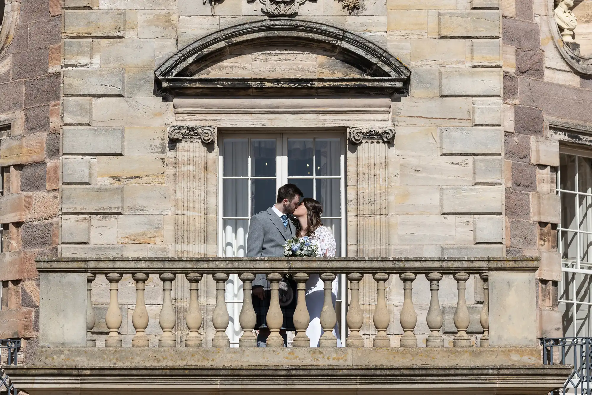 A couple kissing on a balcony of a historic building, surrounded by architectural details and a clear sky.