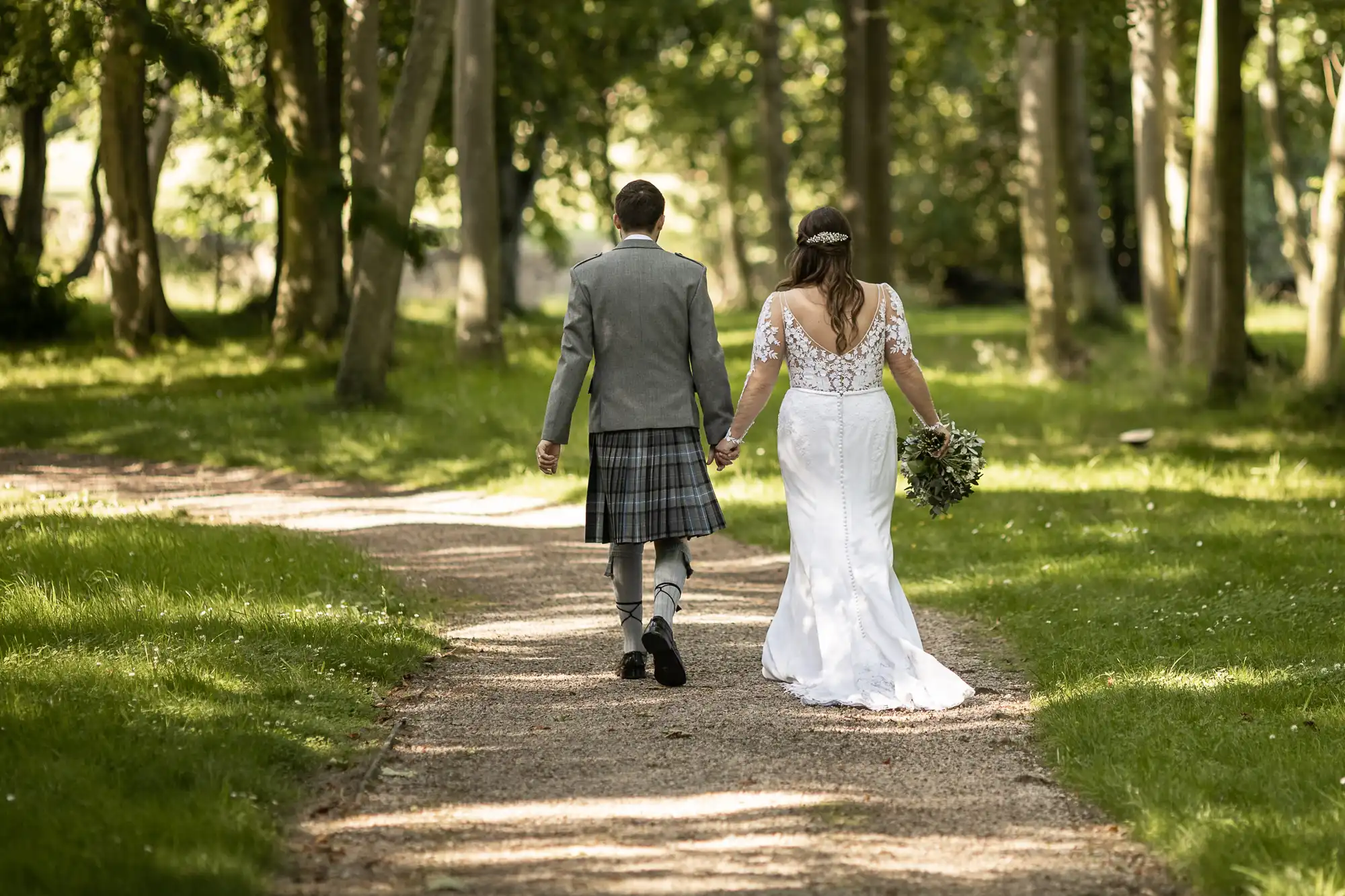 A bride and groom, clad in a white dress and a gray kilt respectively, walk hand in hand down a tree-lined path.