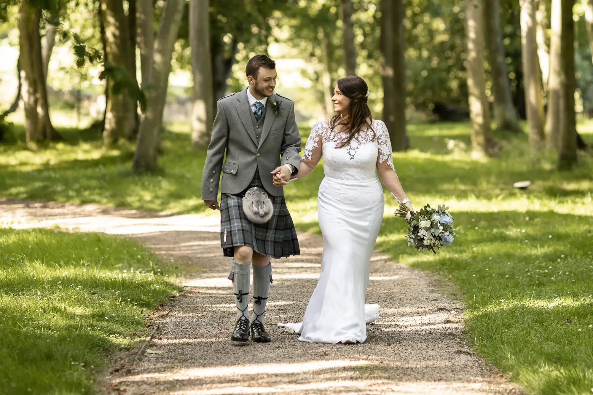 A bride and groom walking hand in hand on a tree-lined path, the groom wearing a kilt and the bride in a white lace gown.
