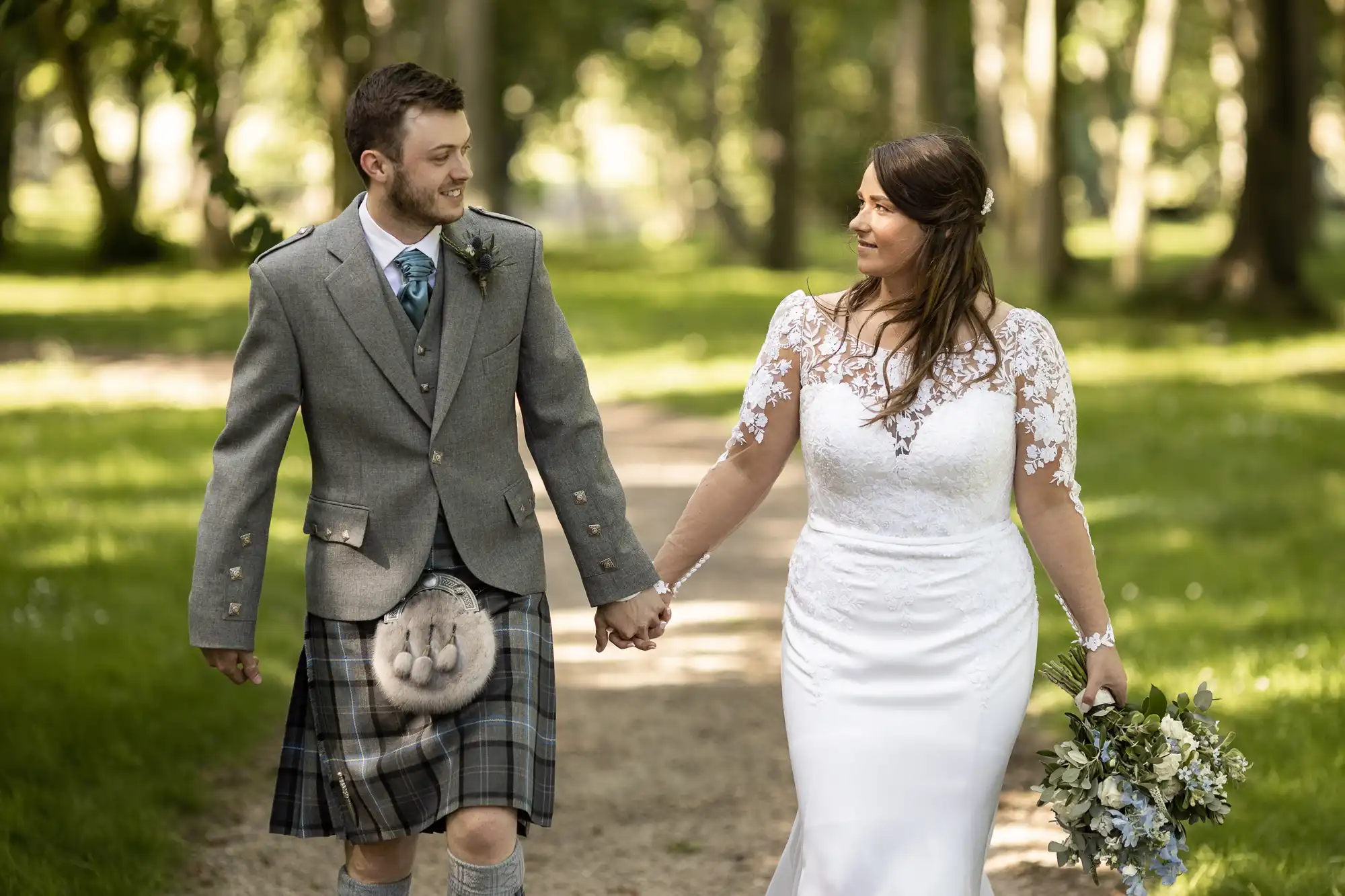 A bride and groom holding hands and smiling at each other as they walk through a sunlit park, the groom wearing a kilt and the bride in a white lace dress.