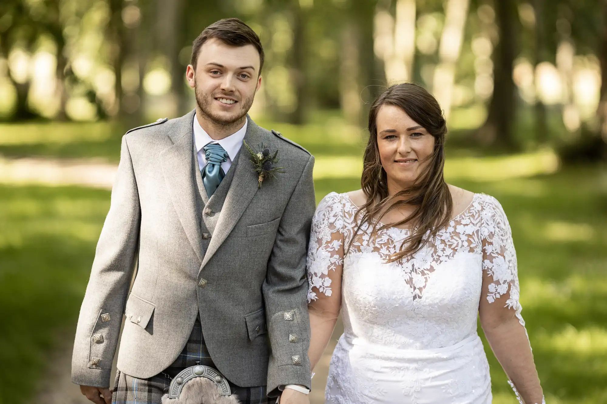 A bride and groom in formal attire smiling outdoors, with the groom wearing a kilt and the bride in a lace dress.