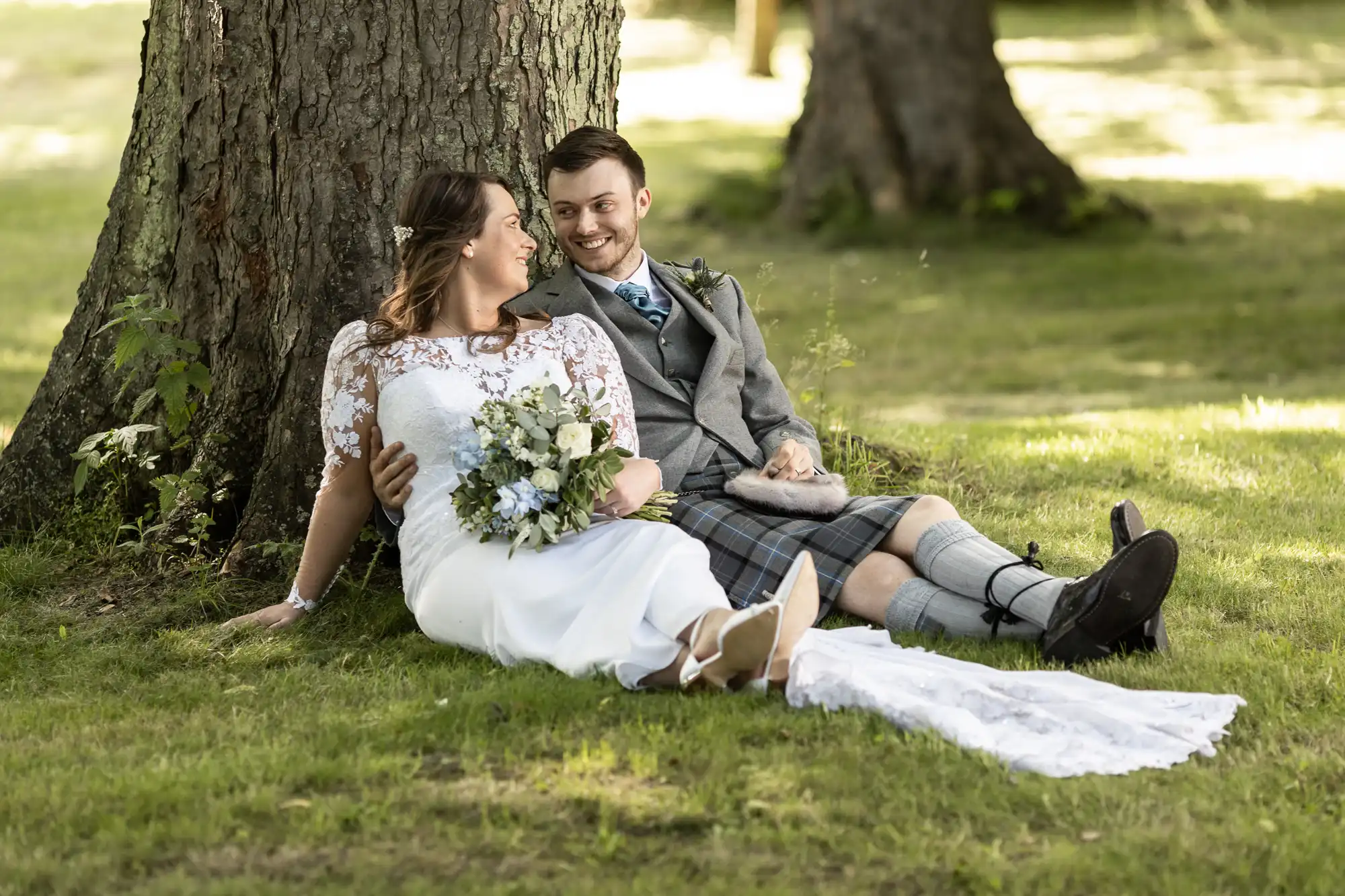 A bride in a white lace dress and a groom in a kilt and tweed jacket sitting under a tree, laughing together, surrounded by grass.
