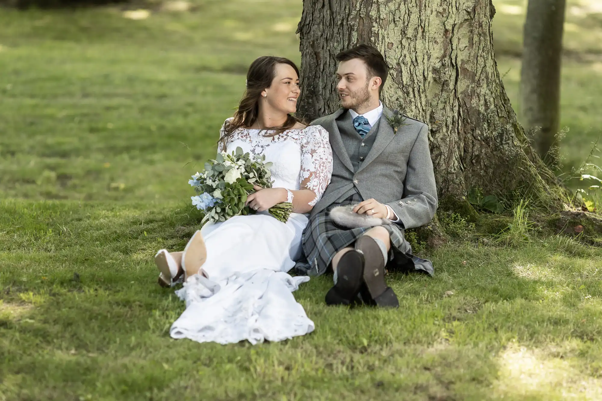 A couple in wedding attire sits under a tree, smiling at each other, the man in a kilt and the woman holding a bouquet.