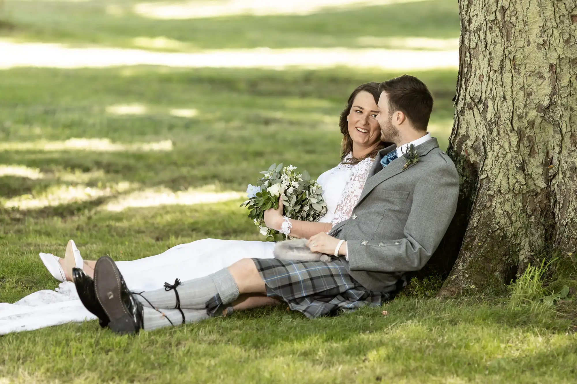 A newlywed couple in wedding attire smiling and sitting under a tree, with the groom wearing a kilt and the bride holding a bouquet.