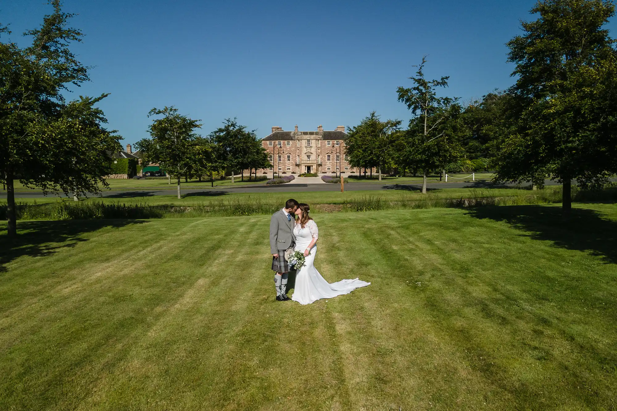 A bride and groom stand holding hands in a grassy field, with a grand estate visible in the background under a clear blue sky.