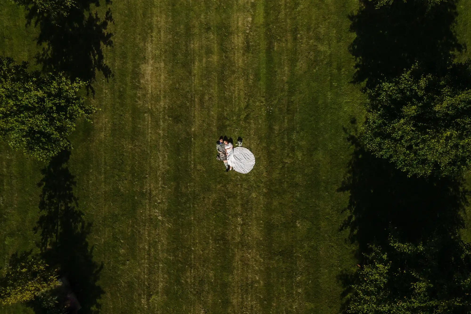 Aerial view of a bride and groom lying on a white round rug in the center of a large green grass field.