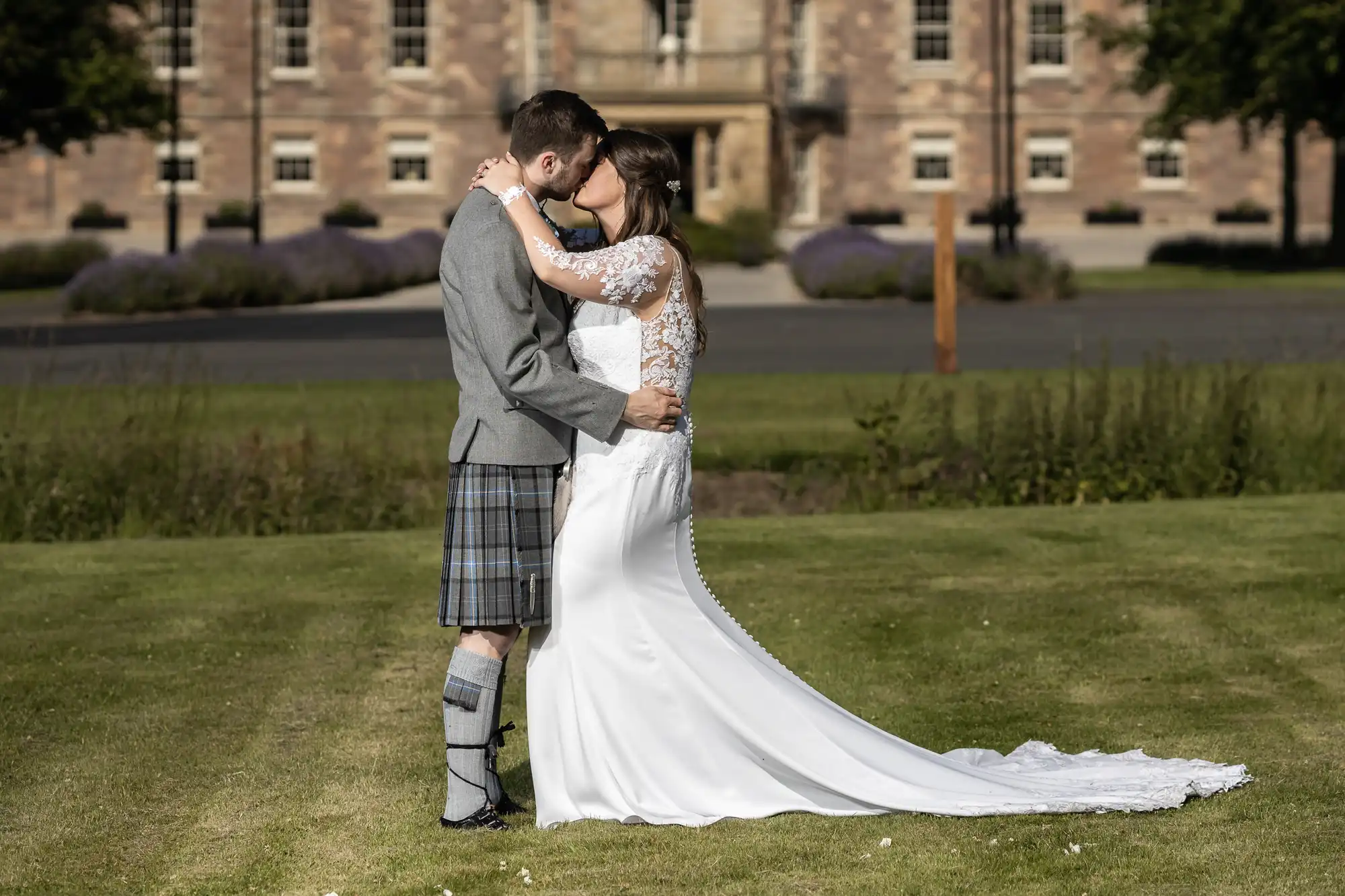 A couple embraces and kisses on a grassy lawn, with the man in a kilt and the woman in a long white wedding dress, in front of an elegant building.