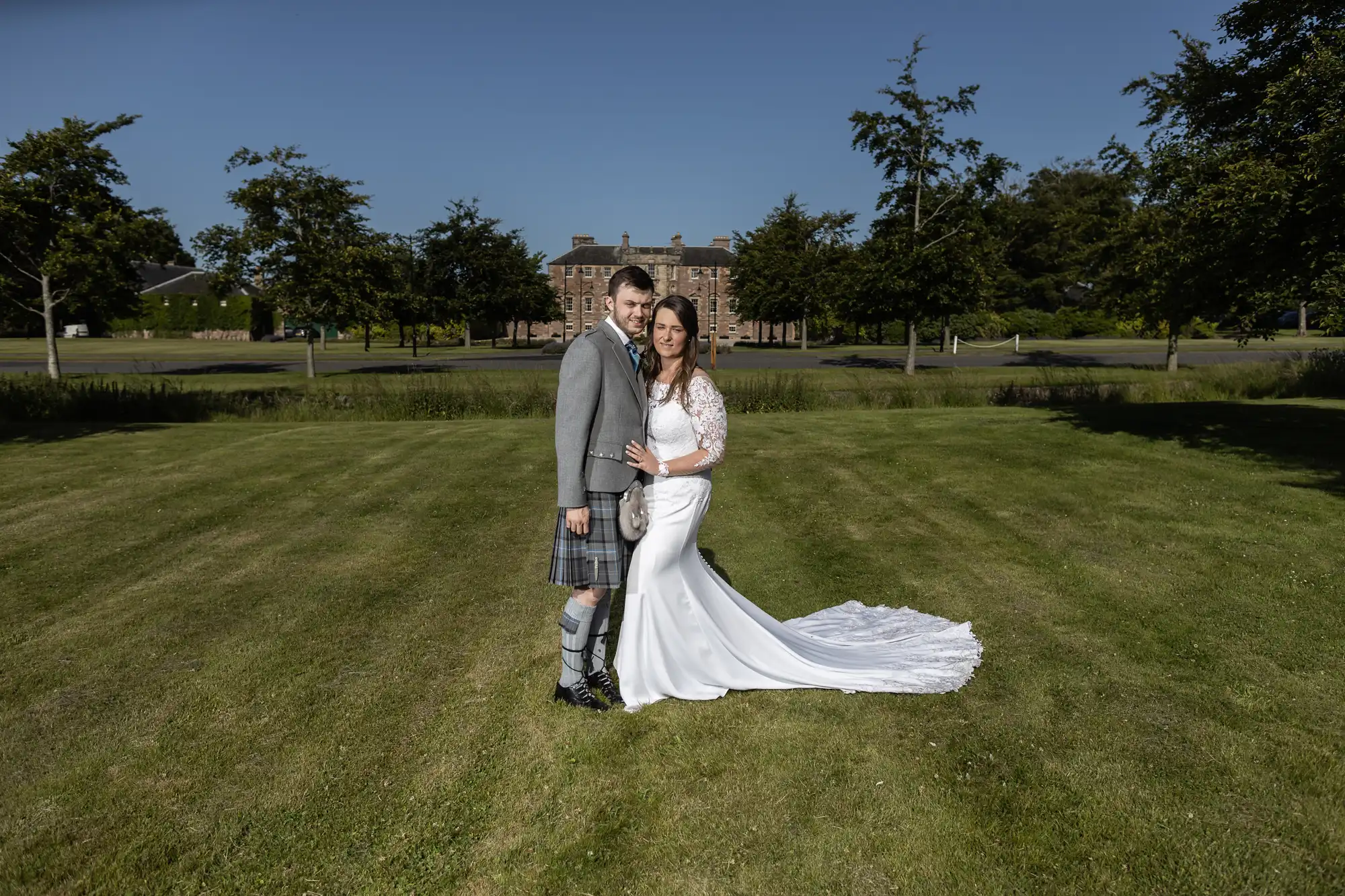 A bride and groom standing on a grassy lawn with a large manor house in the background on a sunny day.