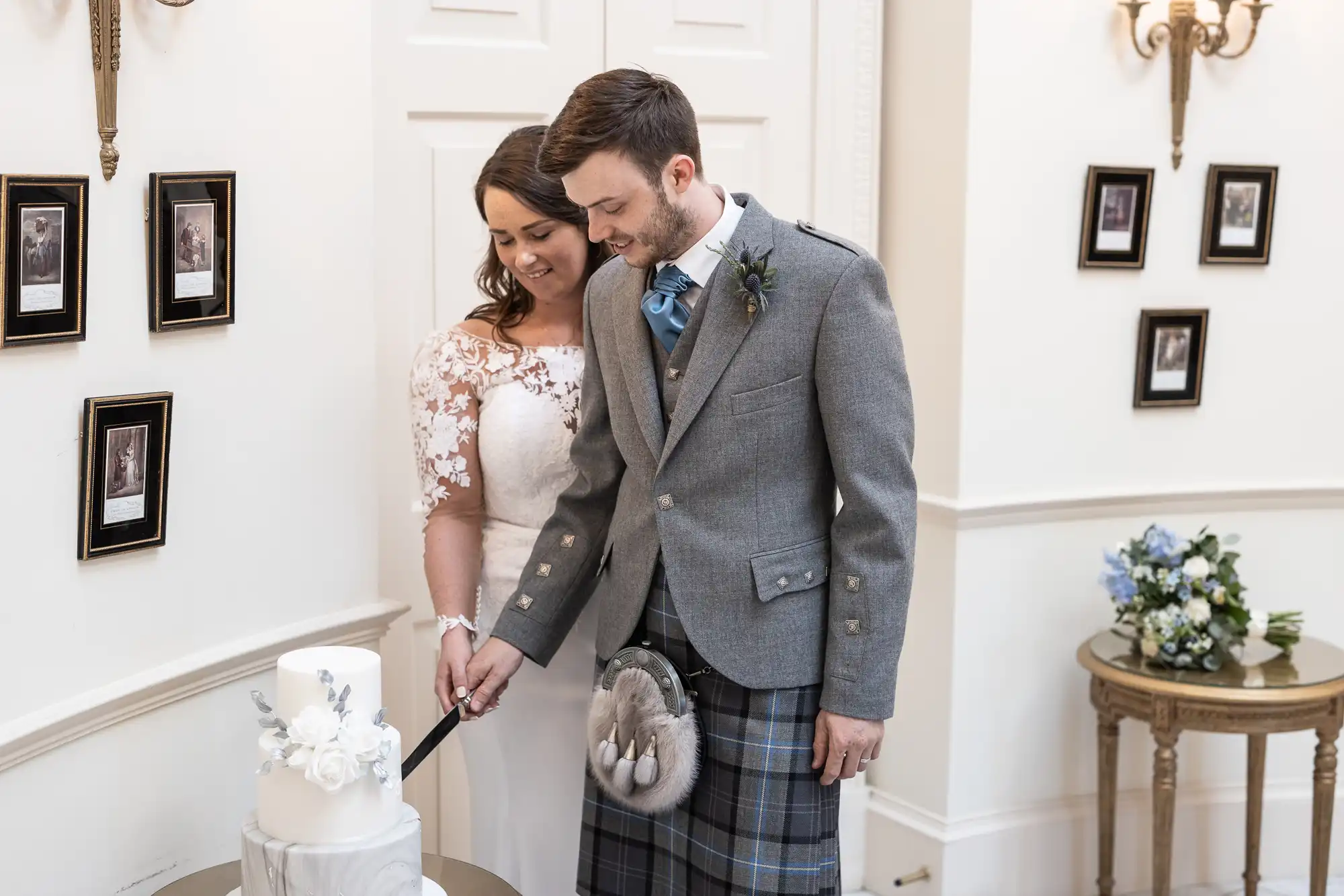 A couple in wedding attire, the man in a kilt, and the woman in a white dress, smiling as they cut a cake together in a room decorated with framed photos.