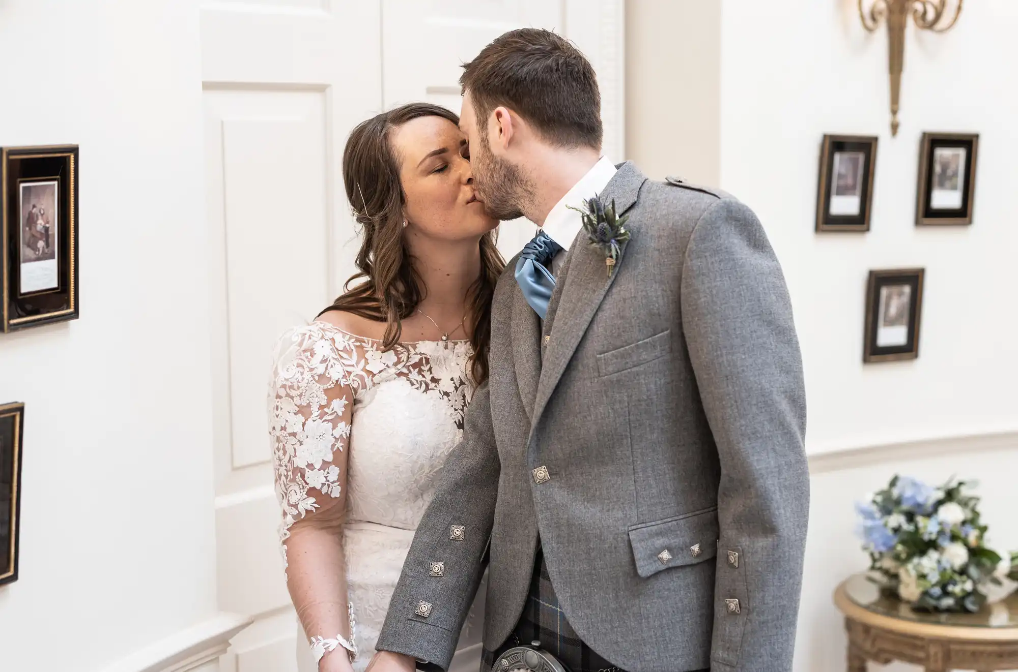 A bride in a white lace dress kisses the groom wearing a gray suit with a blue tie and tartan kilt in an elegant room with framed pictures.