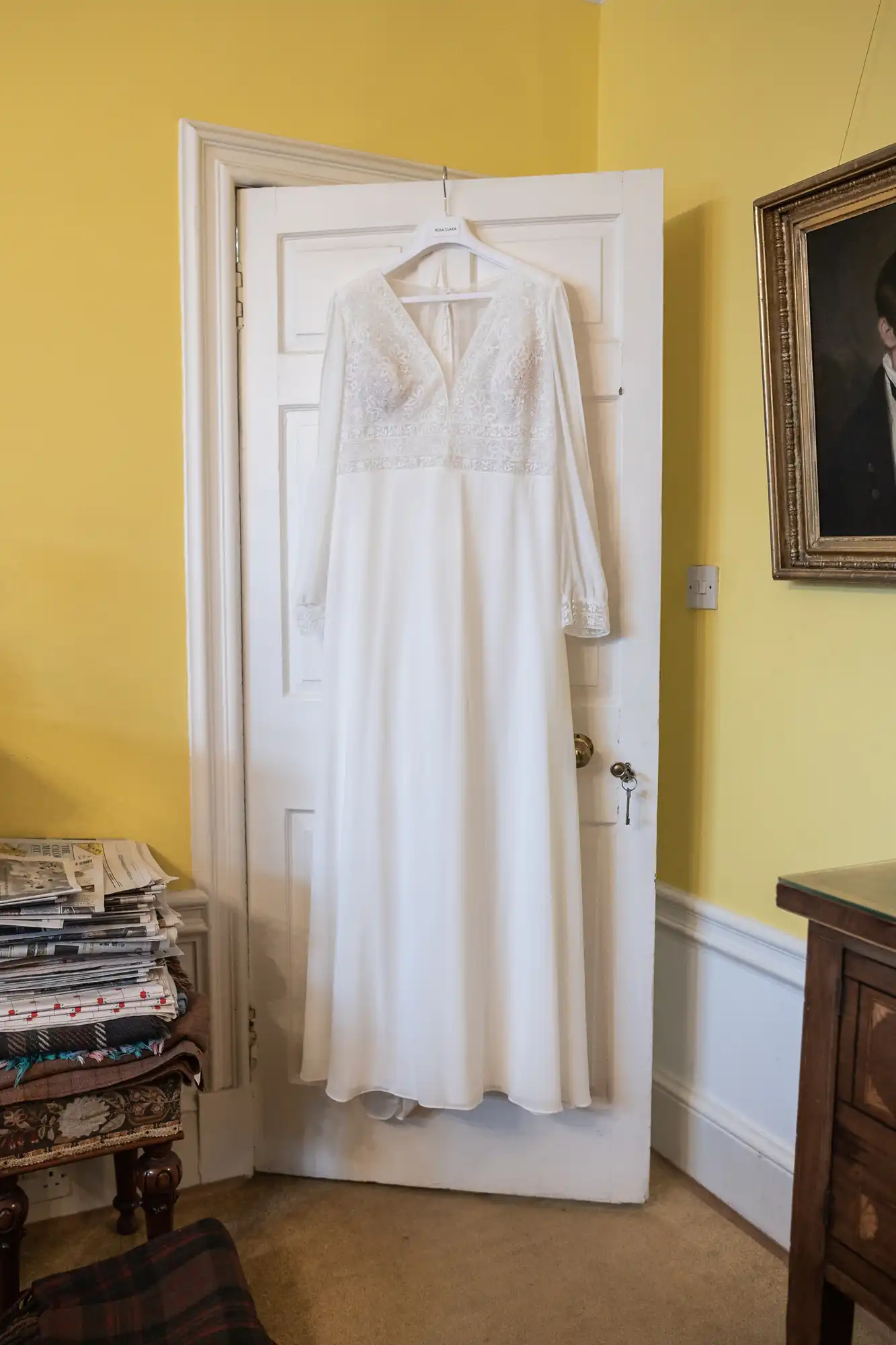 A white long-sleeved bridal gown with lace details hangs on a white door against a yellow wall. A stack of newspapers and framed portrait are visible in the room.