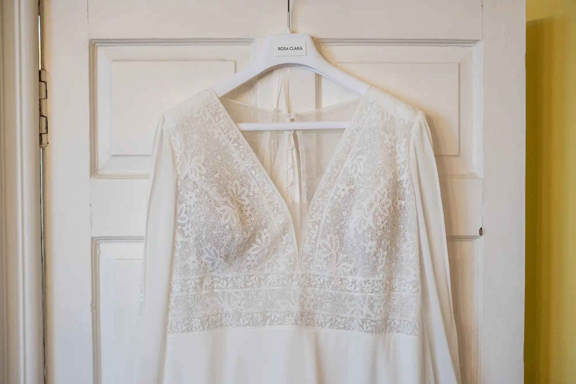 A white lace wedding dress with intricate embroidery, hanging on a white hanger, displayed against a closed white door.