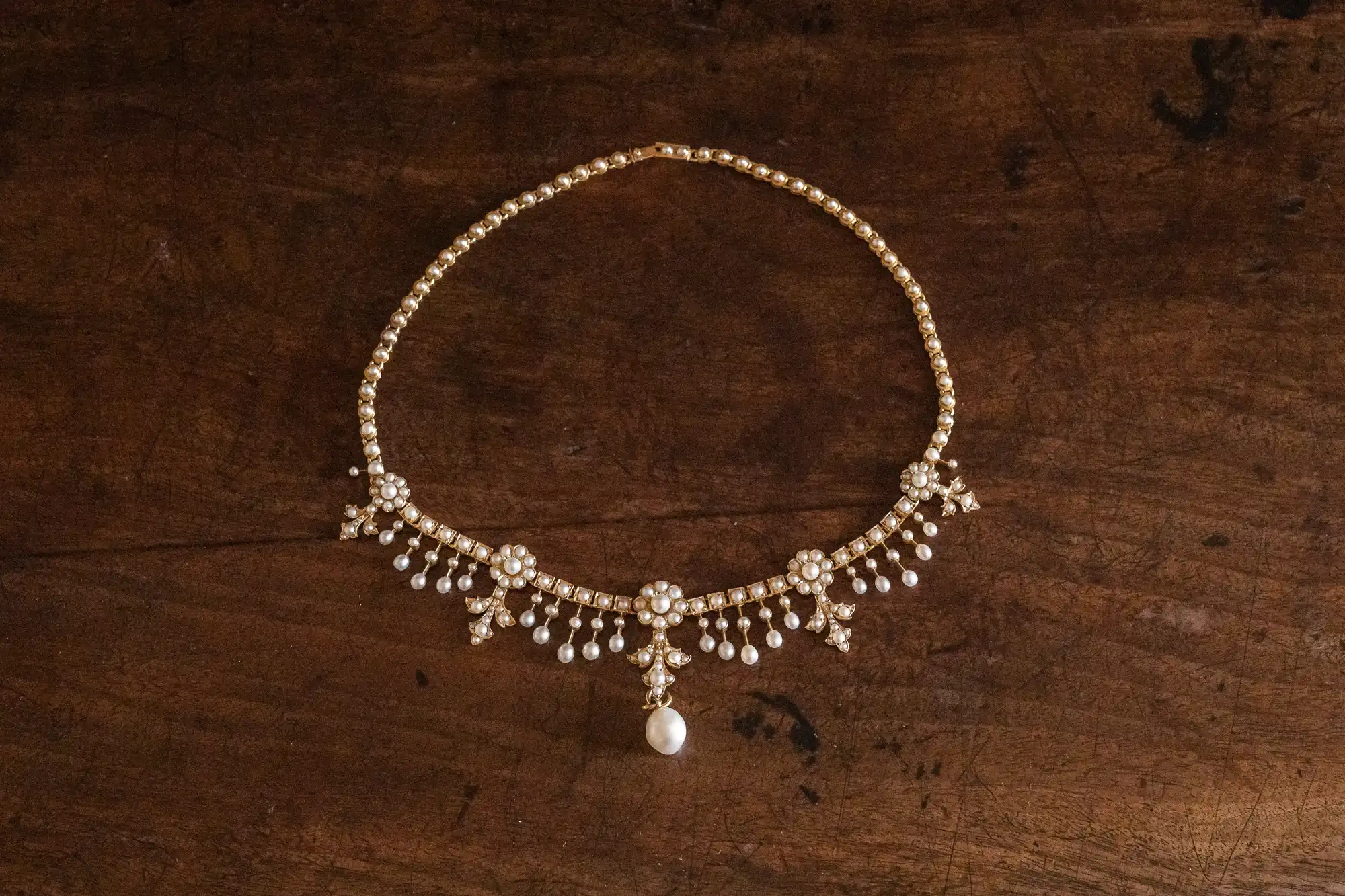 A delicate gold necklace with intricate designs featuring white stones and pearls, arranged on a wooden surface.